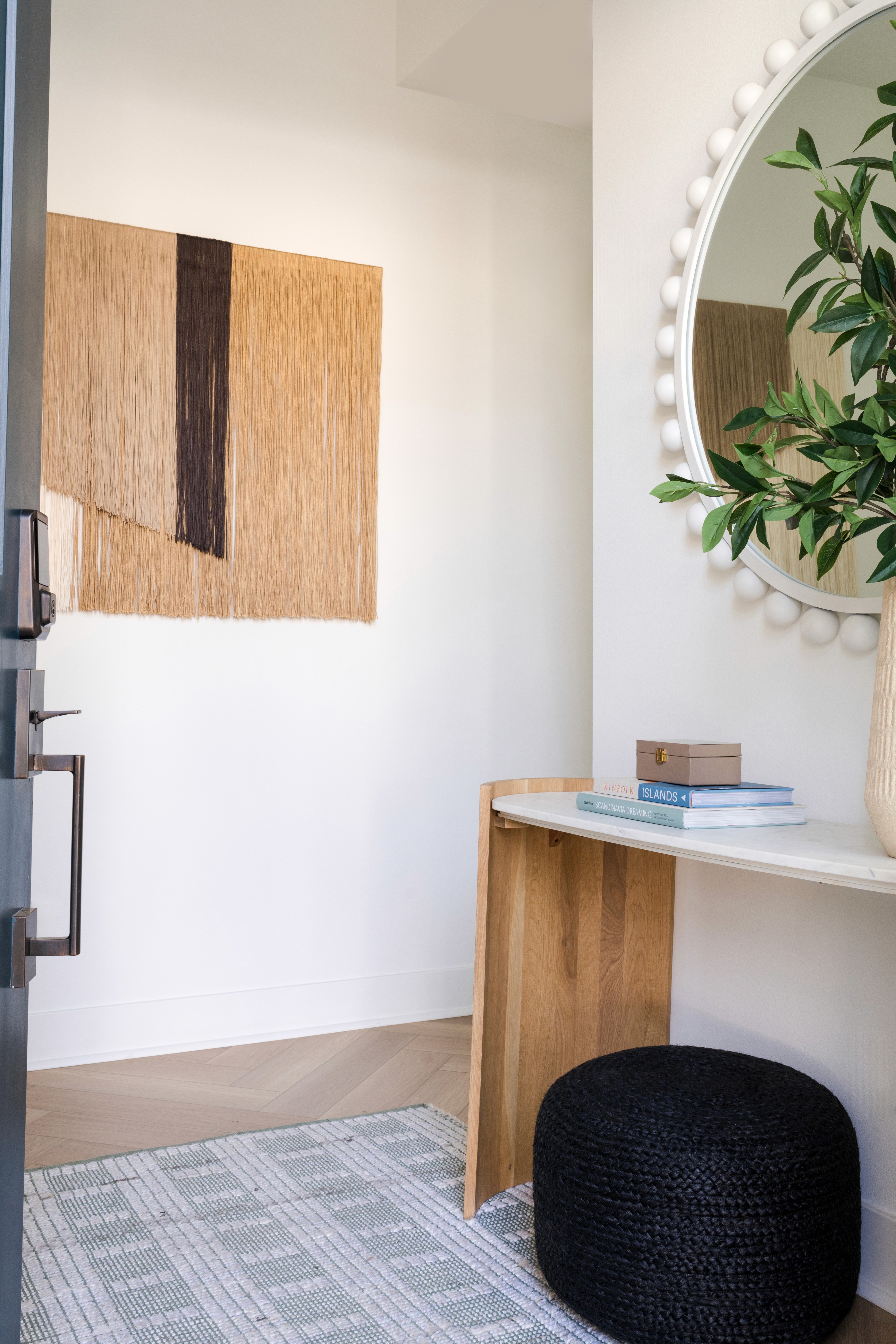Front door view of entryway with cool toned woven rug, modern console table with curved wood legs, circular white mirror, and hanging tapestry by Jubilee Interiors in Arcadia, California