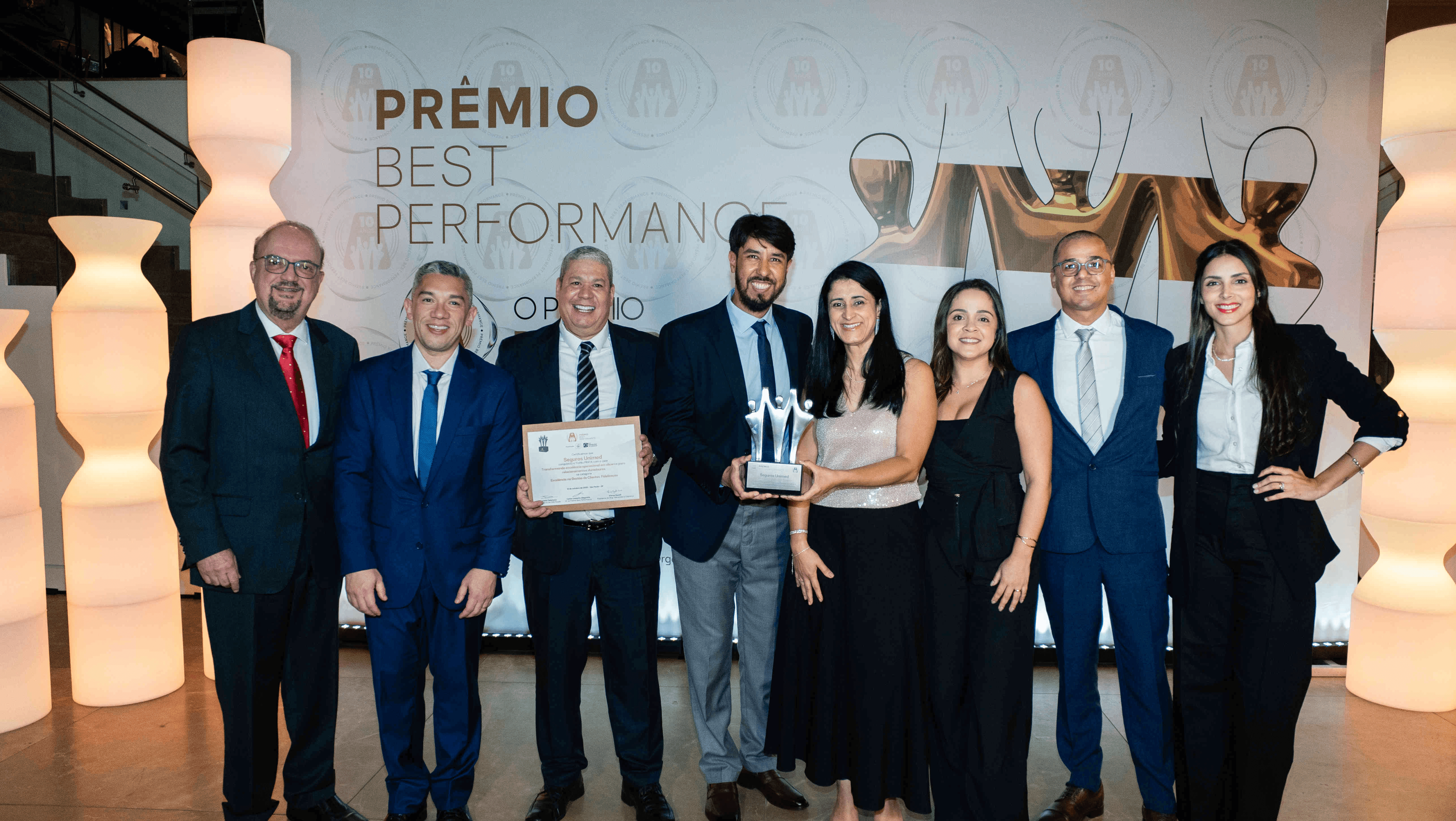 Executivos da Seguros Unimed posando com troféu prata e certificado no palco do Prêmio Best Performance 2025