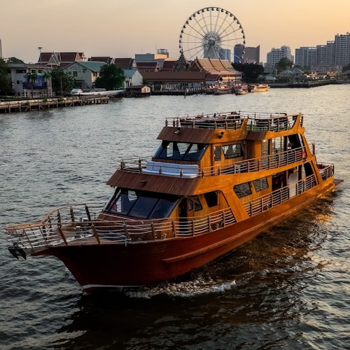 O barcă din lemn navighează pe un râu lângă un oraș unde o roată panoramică și clădiri sunt vizibile pe fundal în timpul apusului.