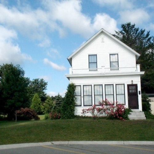 บ้านสีขาวสองชั้นพร้อมระเบียง ล้อมรอบด้วยสนามหญ้าสีเขียว พุ่มไม้และต้นไม้ ใต้ท้องฟ้าที่มีเมฆบางส่วน