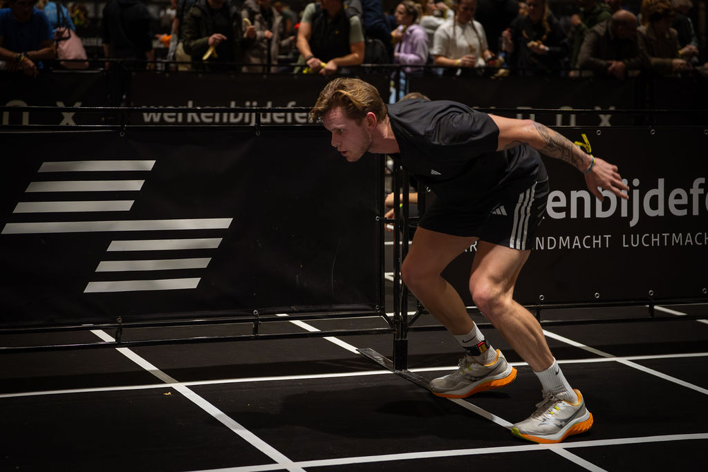 A young person with curly hair smiles at the camera while sitting in front of a laptop against a dark background.