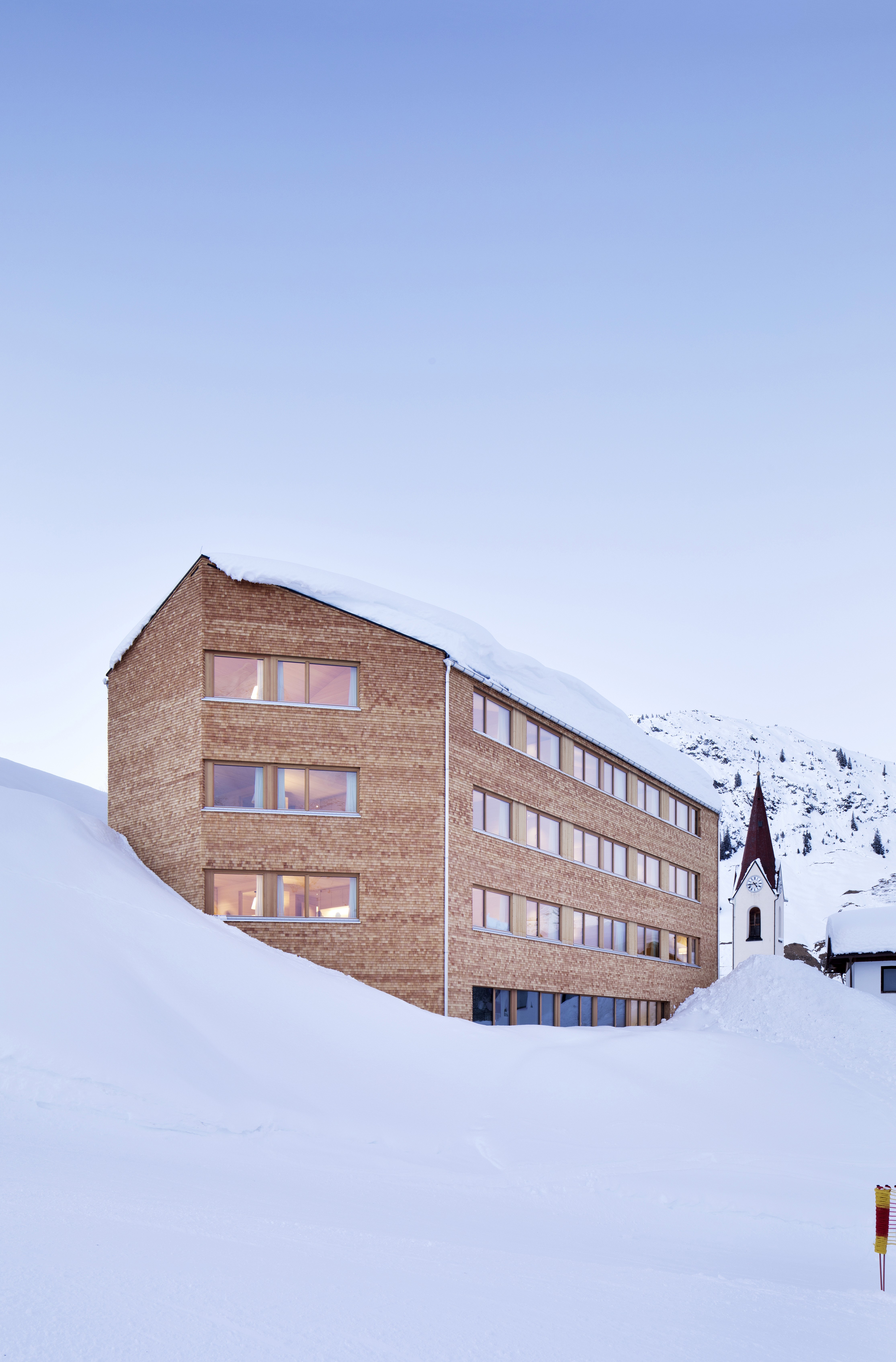 Contemporary alpine building with a wooden façade, large windows glowing from inside, and deep winter snow surrounding it, with a small church tower beside it.