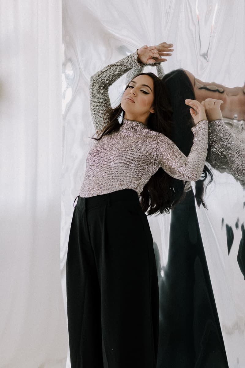 A model standing in front of a reflective surface, wearing black trousers and a silver top, striking a pose for a creative Mylar shoot at Revelator Studio, a natural light studio in Shreveport.