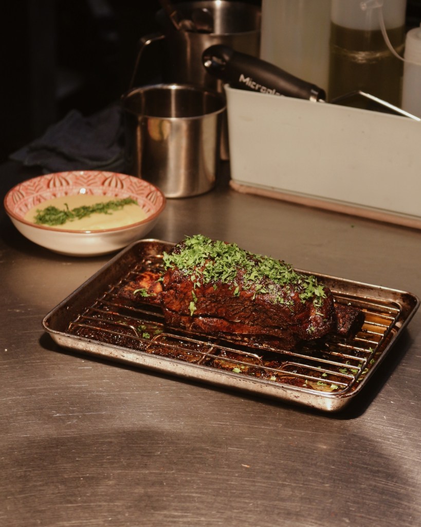A beautifully cooked piece of meat garnished with fresh herbs, resting on a wire rack over a tray, with a bowl of sauce in the background.