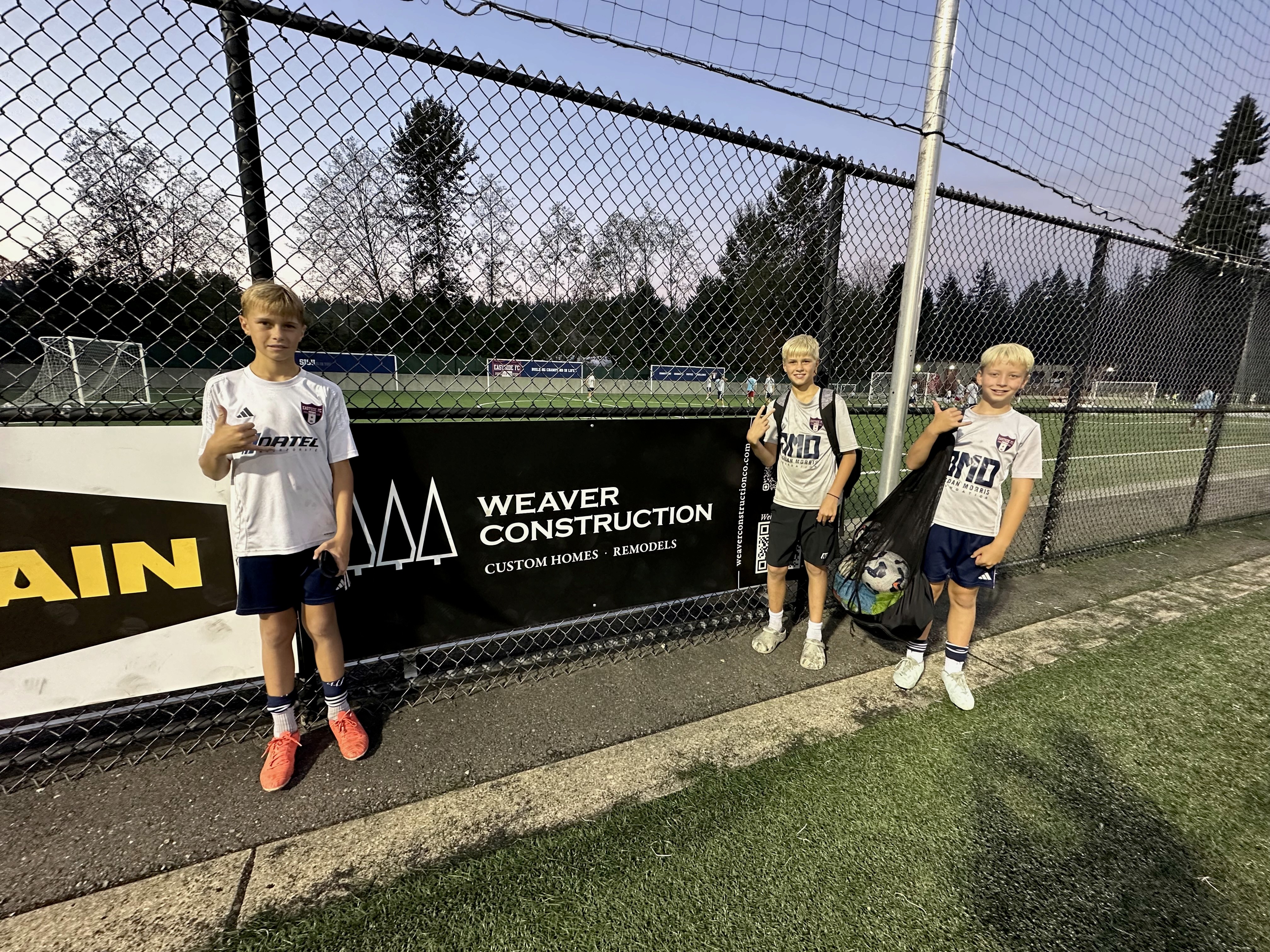 An image of three youth soccer players at a training ground next to a sponsor sign of Weaver Construction