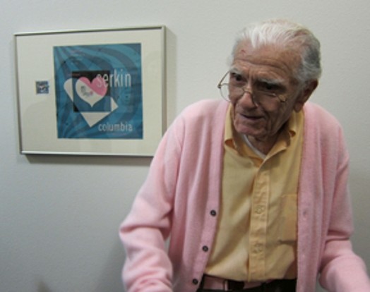 A photo of an old man wearing glasses, a yellow shirt and a pink blouse sitting in front of a blue printing with frame that shows a white and pink heart.