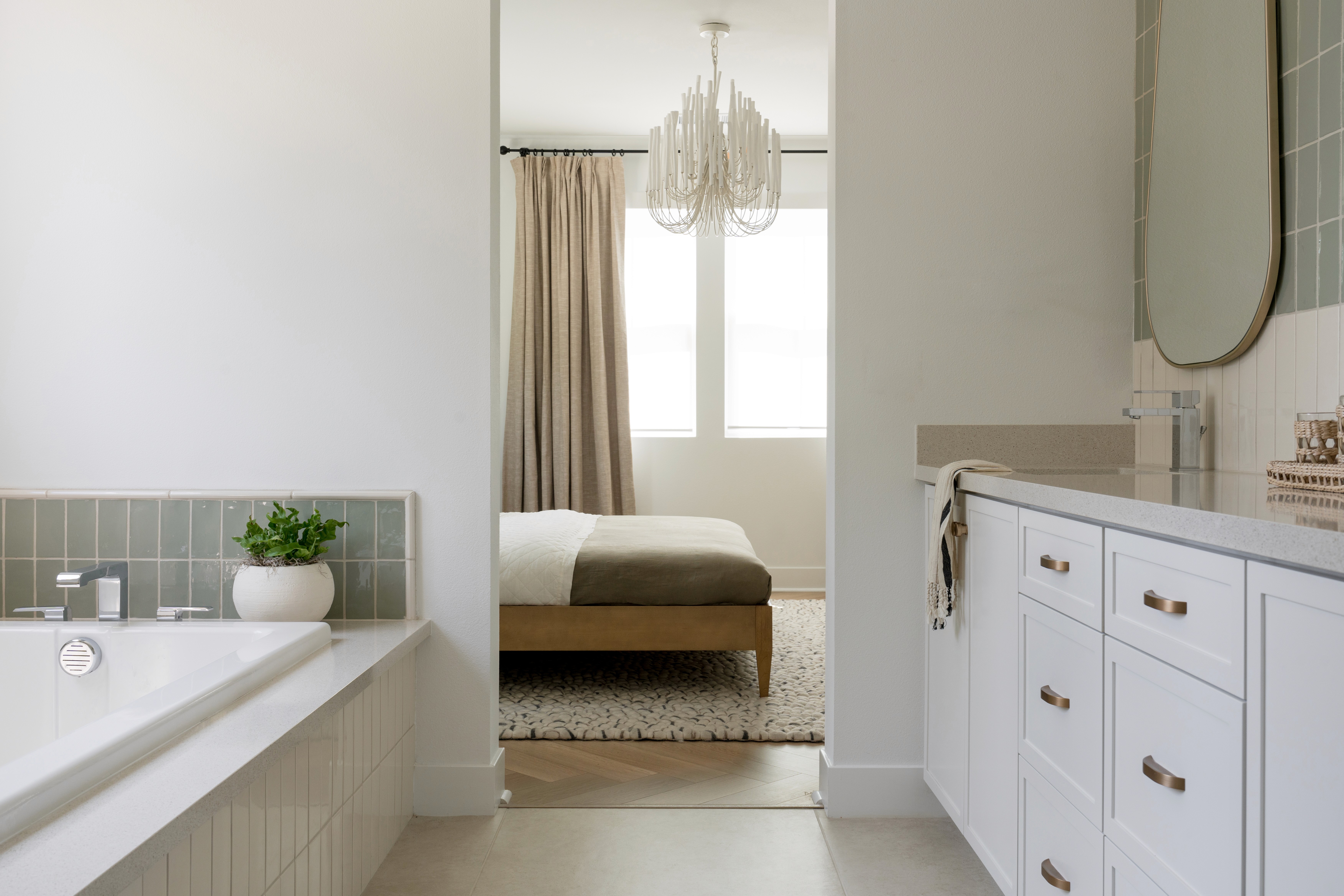 White tiled, rectangular bathtub with sage green tile surrounding tub, white cabinetry and drawers with brass finishes, and oval vanity mirror by Jubilee Interiors in Arcadia, California