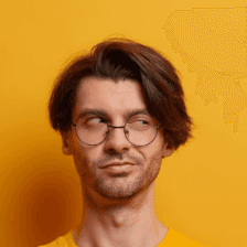 A man with brown hair and glasses looks sideways with a curious expression, standing against a vibrant yellow background while wearing a matching yellow shirt.