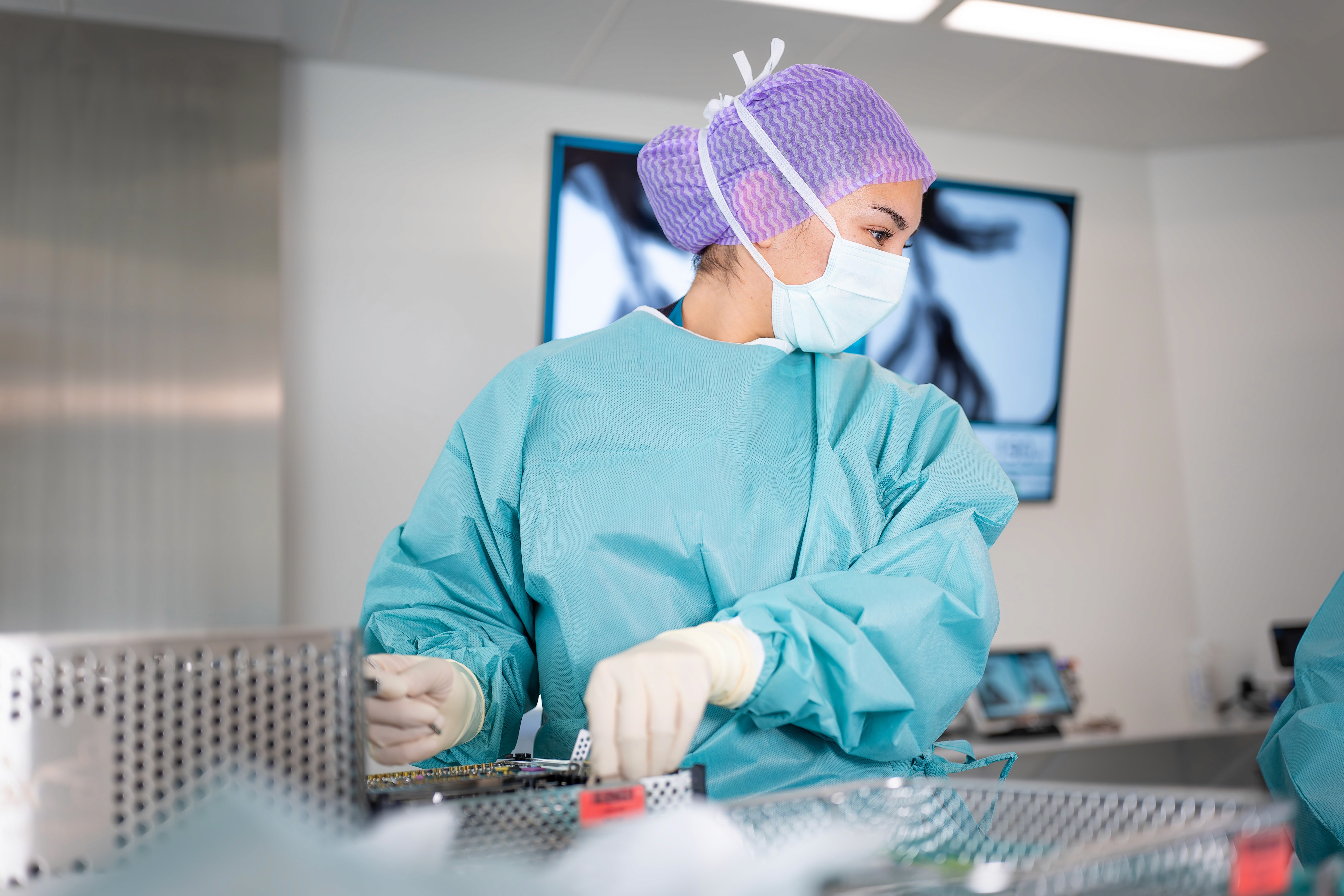 Ein medizinische Fachkraft in blauem OP-Kittel, lila Kappe und Maske bereitet in einem hellen Operationssaal die Instrumente vor, während im Hintergrund medizinische Geräte und ein Monitor zu sehen sind.
