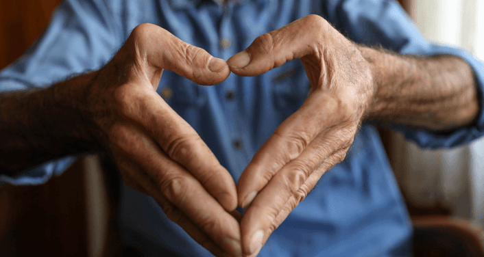 Close-up of an elderly person's weathered hands forming a heart shape, with a blue shirt visible in the soft-focus background.