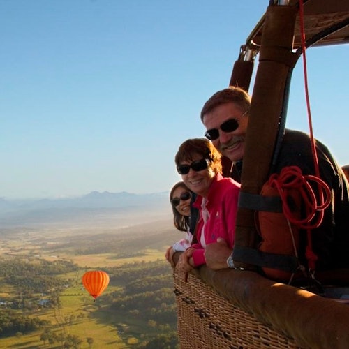 Drie mensen met zonnebrillen glimlachen vanuit een heteluchtballonmand, terwijl een andere oranje ballon zichtbaar is boven een schilderachtig landschap beneden.