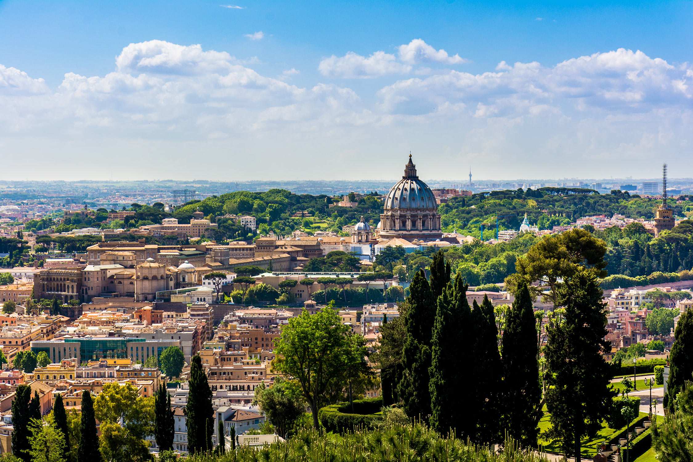 Panoramic view of Rome's cityscape featuring St. Peter's Basilica dome under a blue sky with white clouds.