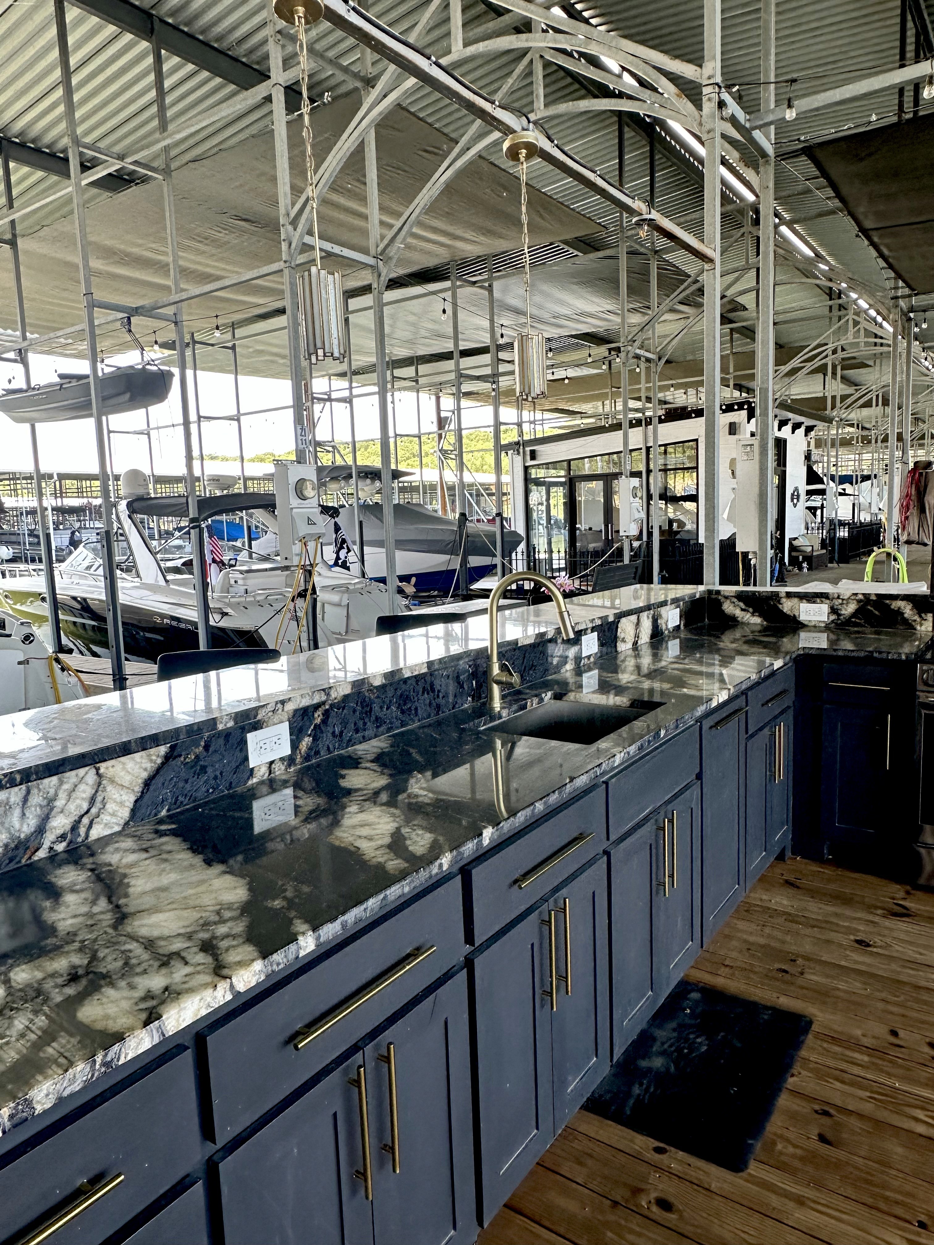 Luxurious modern kitchen with elegant dark blue cabinetry and a polished marble countertop, featuring sleek gold hardware, set within a spacious boat dock with several boats in the background.