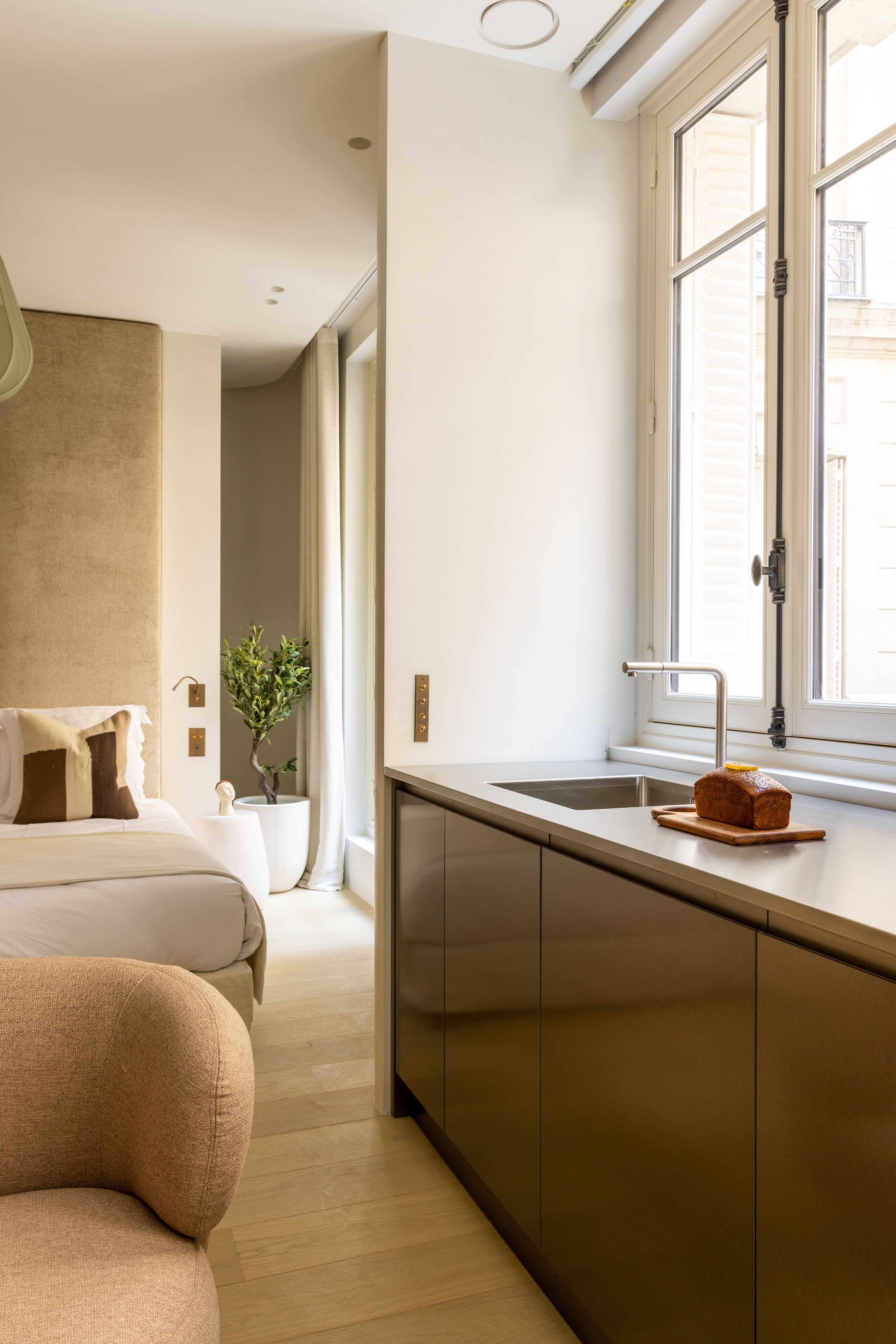Vue transversale kitchenette bronze avec evier, pain brioché, et chambre avec tete de lit velours en arriere-plan