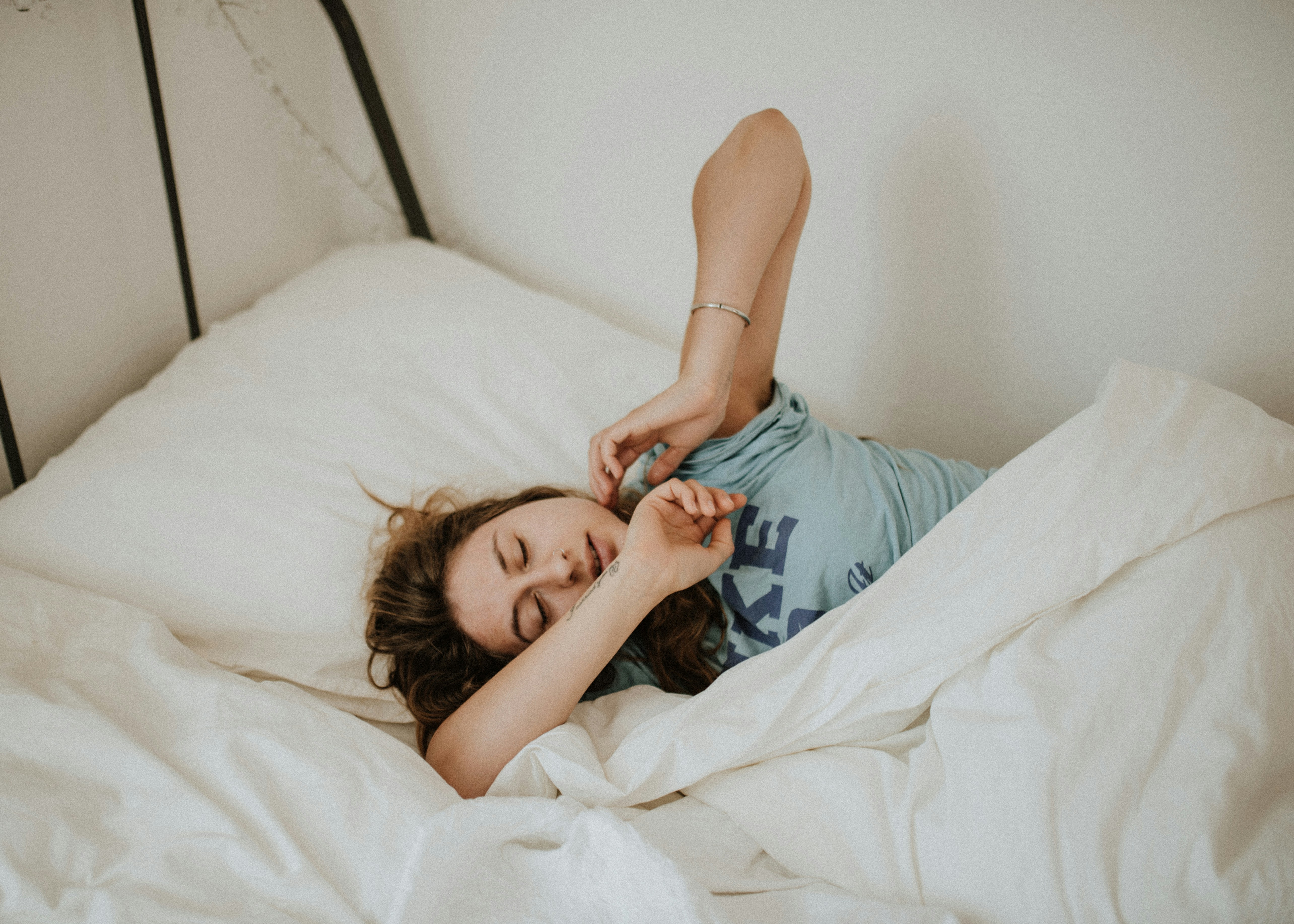 A woman lies in bed, stretching as she wakes up. Only a part of the bed can be seen, with the background wall painted white. 