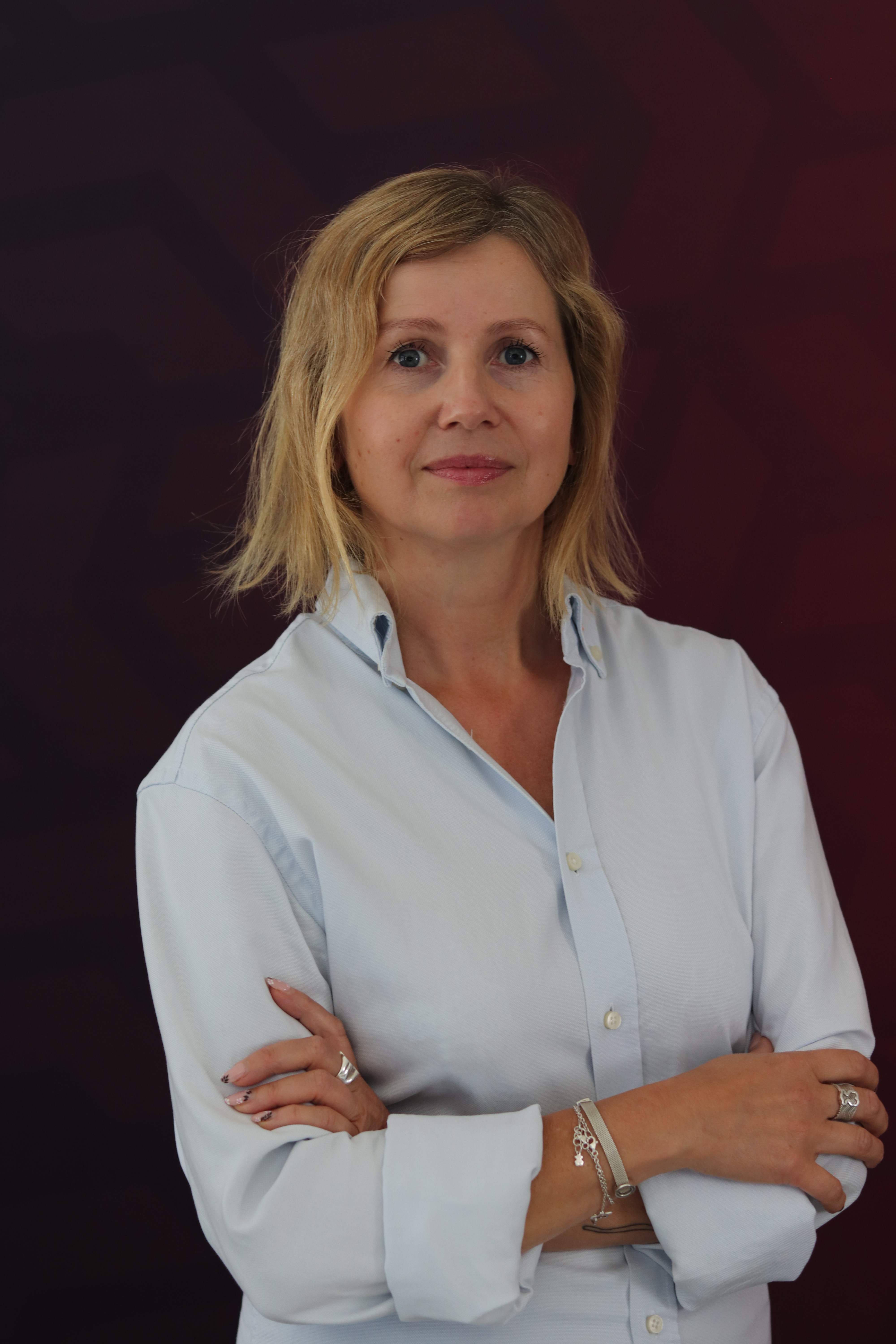 Kobieta w jasnej koszuli z założonymi rękami pozuje na tle bordowej ściany, patrząc z lekkim uśmiechem w stronę kamery.