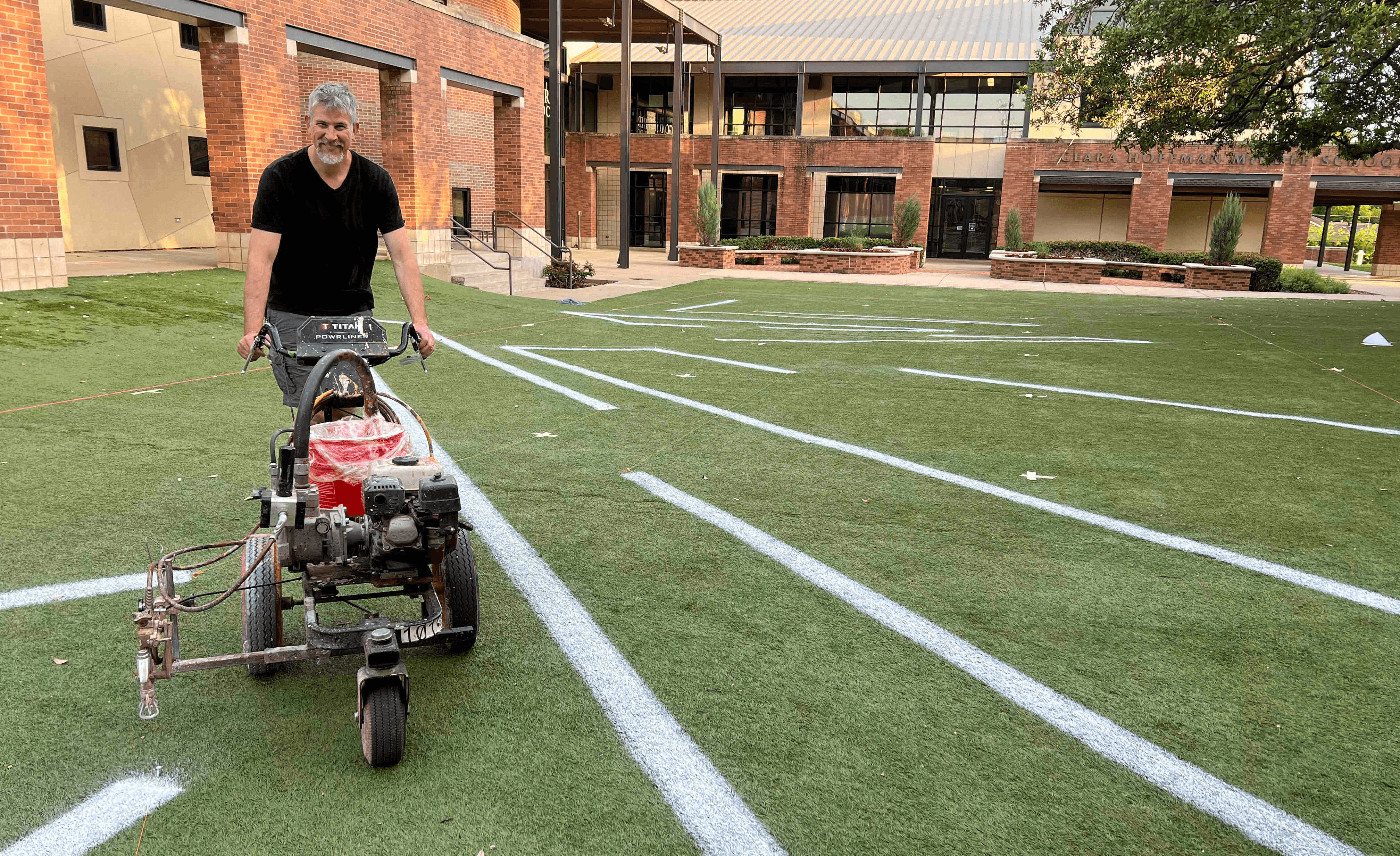 A person using a machine to mark lines on a green turf field, with a building in the background.