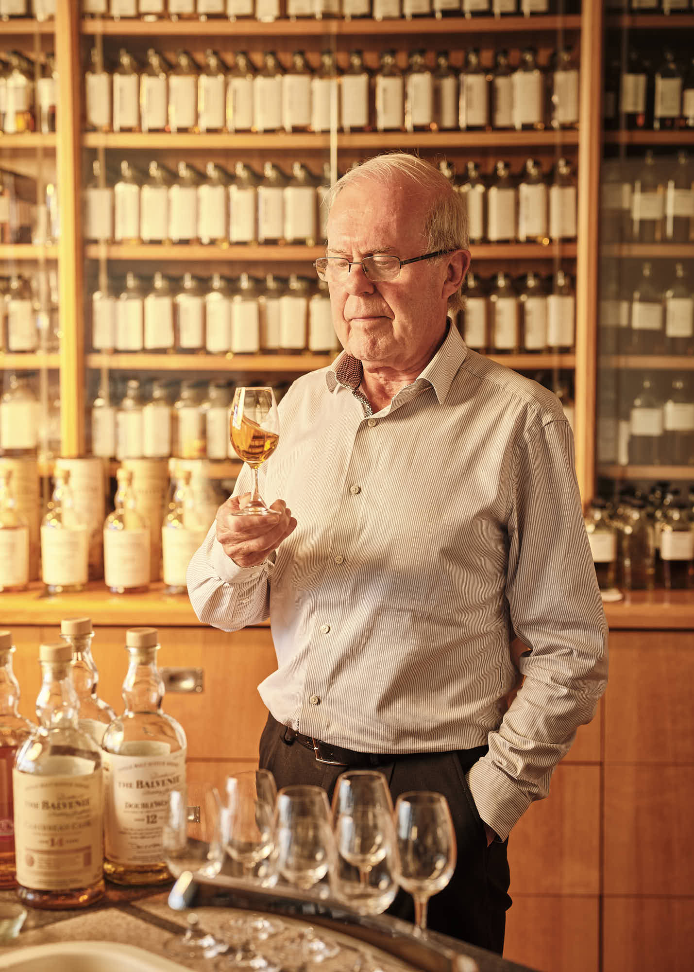 A person stands in a nosing room, examining a dram of whiskey with shelves of bottles in the background.