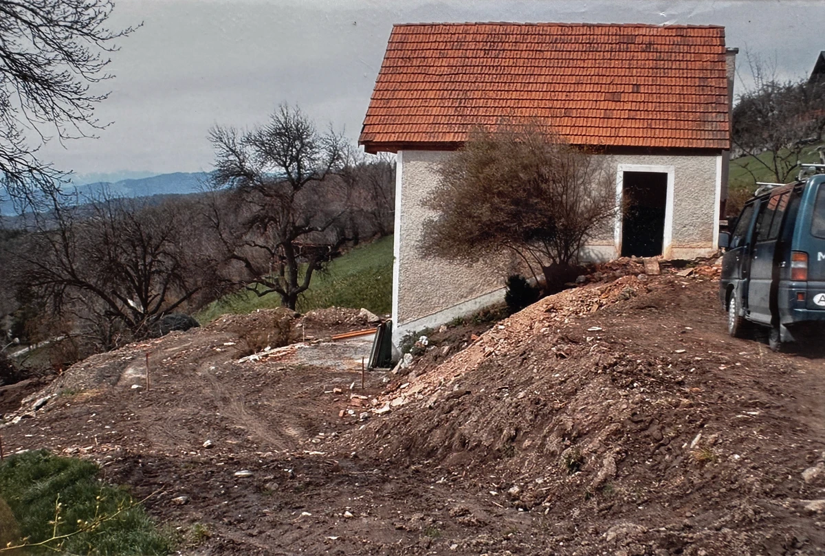 Kleines Haus mit rotem Ziegeldach vor dem Umbau, umgeben von frisch ausgehobenem Erdreich auf der Baustelle mit Blick ins Tal.