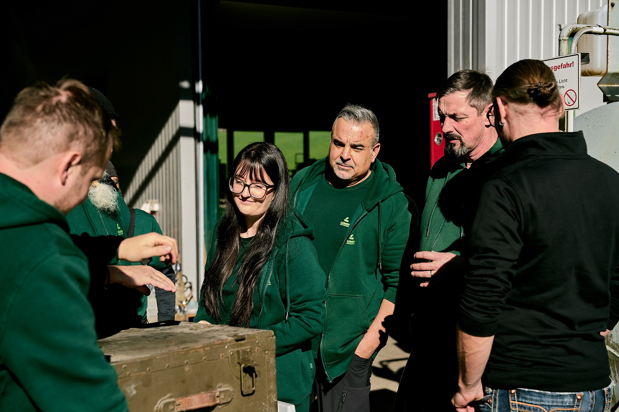 Gruppe von Mitarbeitern in grünen Jacken, die sich um eine alte Truhe versammeln. Sie schauen konzentriert.