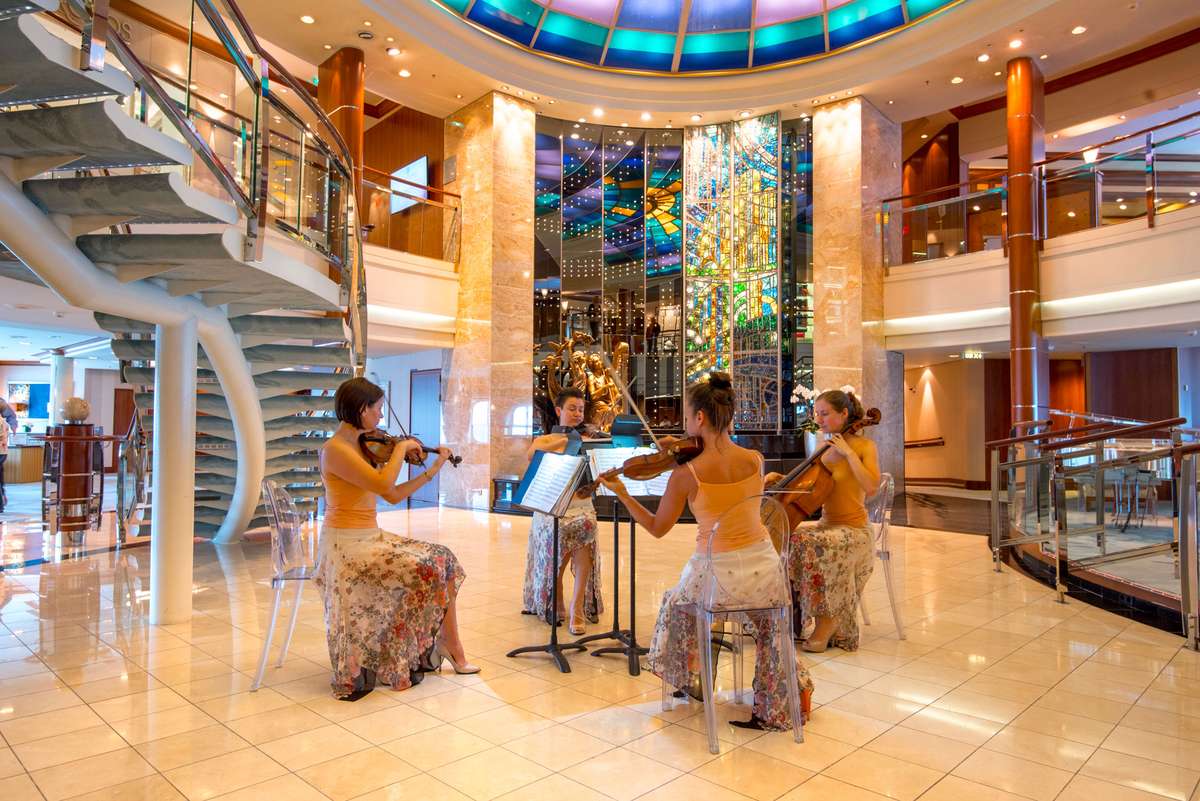 Three women in swimwear sit at a bar and on stools in a cruise ship atrium with a large stained-glass mural.