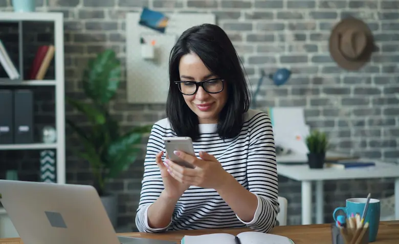 A woman sitting at a desk smiling while using her phone, with a laptop and home office setting in the background.