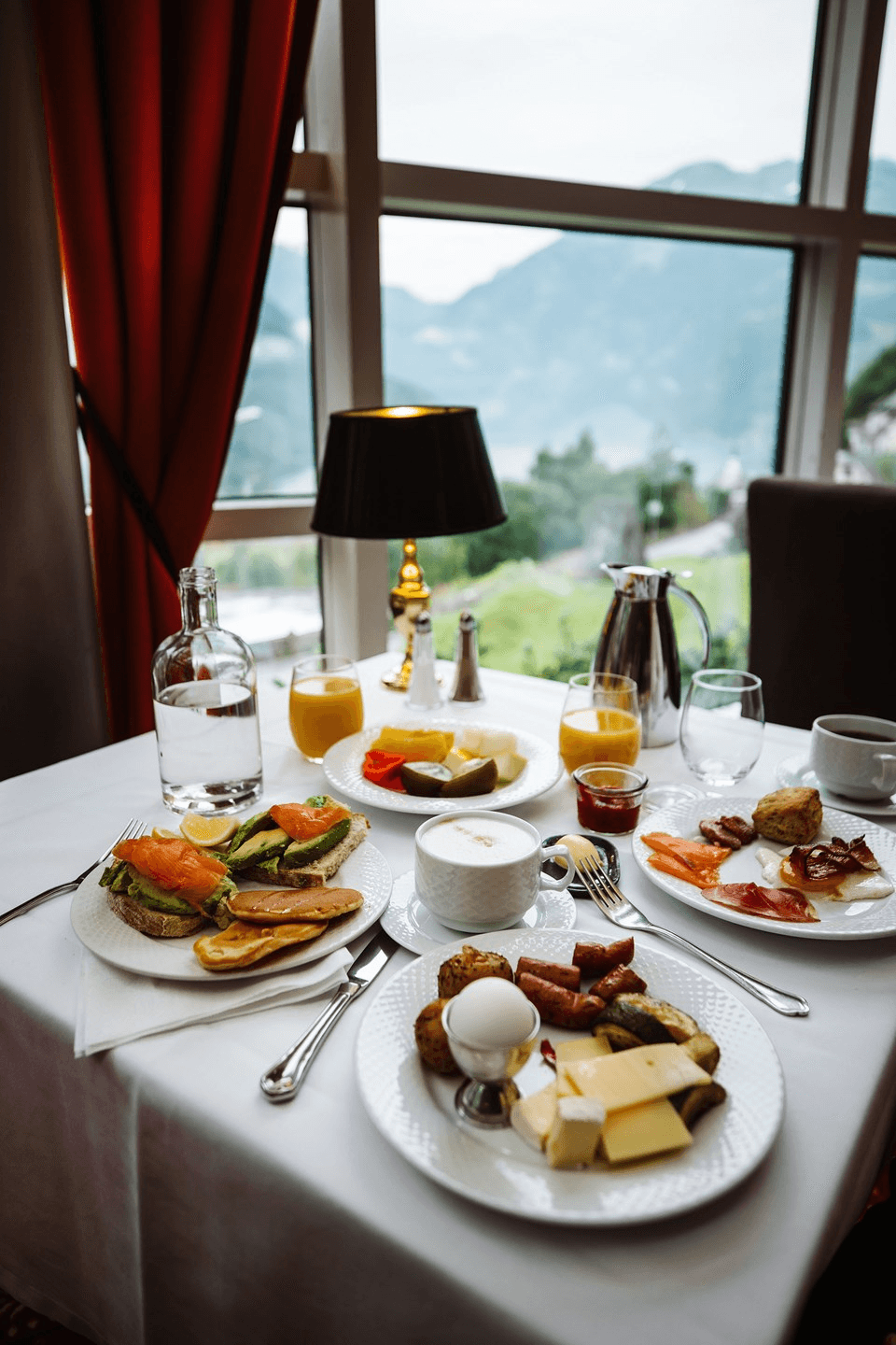 A beautifully set breakfast table next to a window with a scenic mountain view, featuring plates of assorted foods including sliced salmon, cheeses, boiled eggs, pastries, and glasses of orange juice.