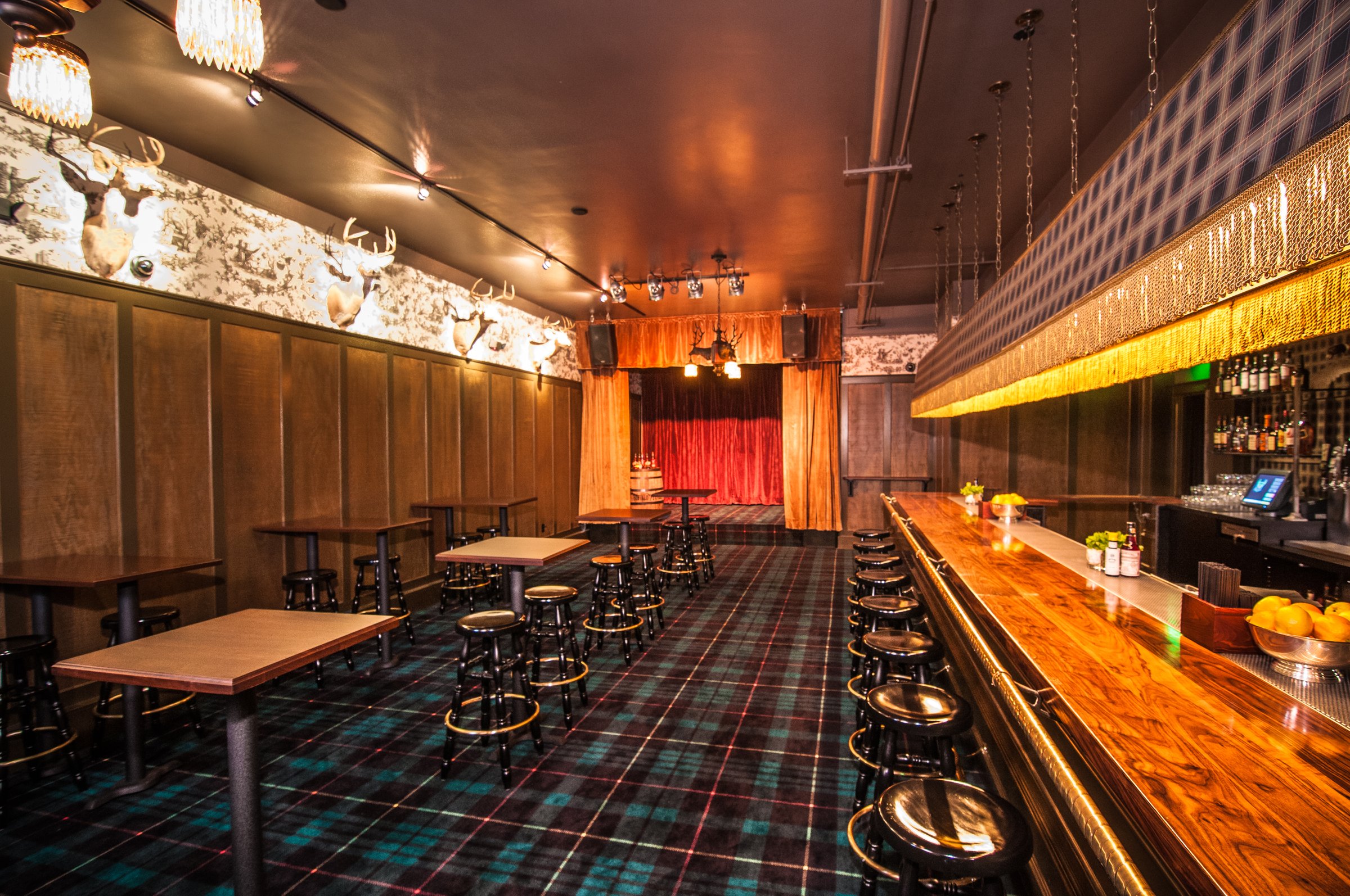 Long timber bar and rows of black stools beside tartan flooring, with a red curtain at the far end.