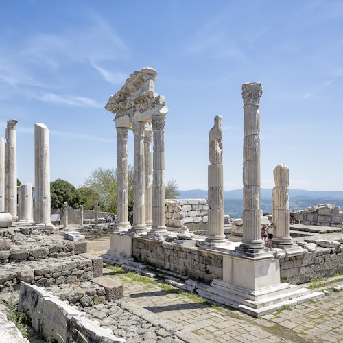 Ancient Greek ruins with tall, partially-intact stone columns under a clear blue sky. People walk among the ruins in the background.