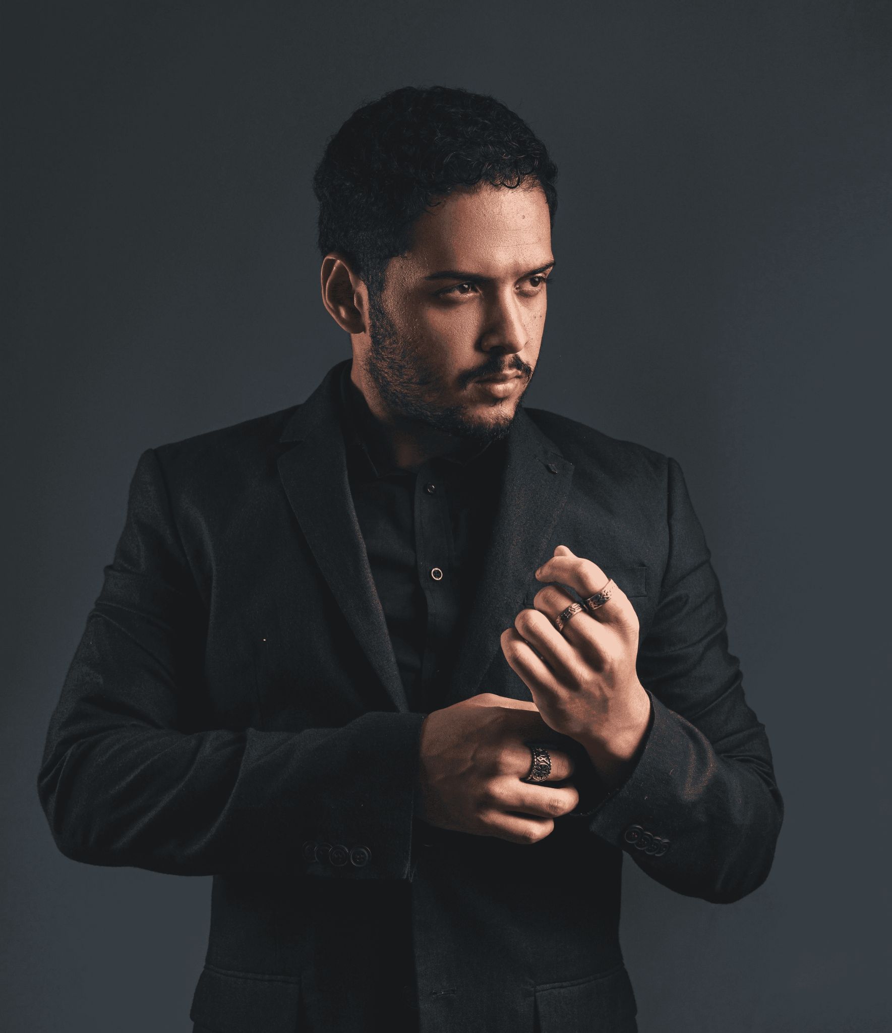 A man in a dark suit adjusts his sleeve, standing against a muted, dark background, exuding an air of sophistication and elegance.