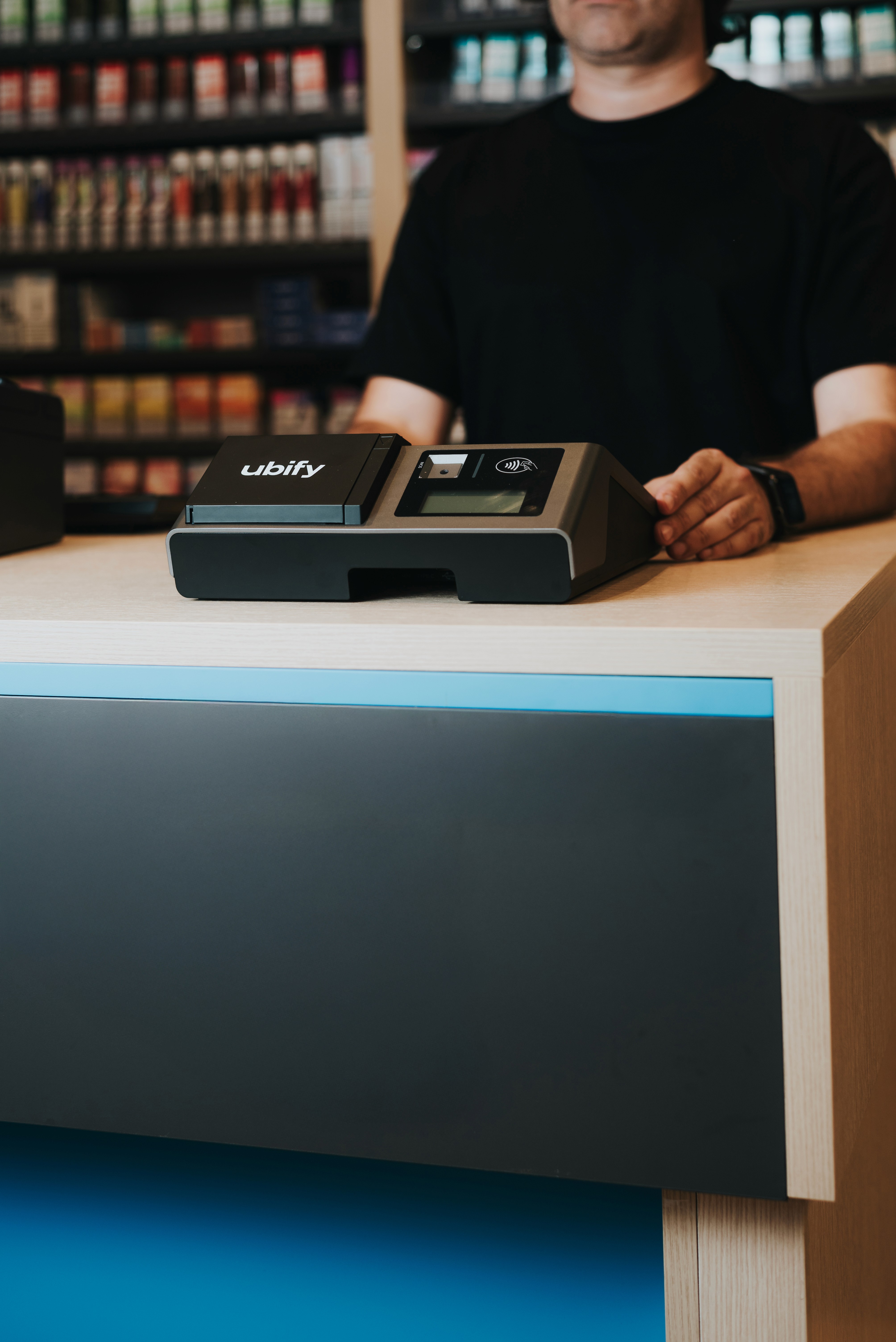 Commerciante dietro al bancone con Cassa Ubify in primo piano, pronta all’uso in un negozio ben fornito.