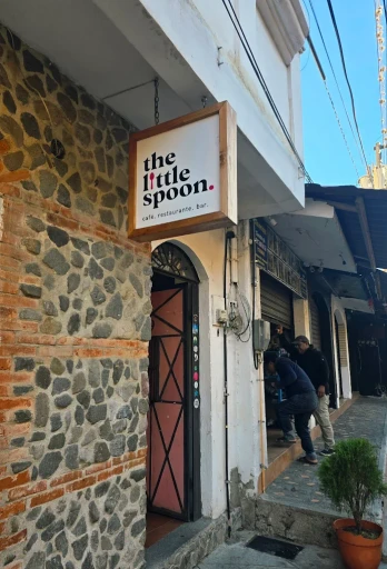 Exterior of a small café with a hanging sign reading “the little spoon – café, restaurante, bar,” set against a stone and brick wall, with two people entering from a narrow sidewalk.