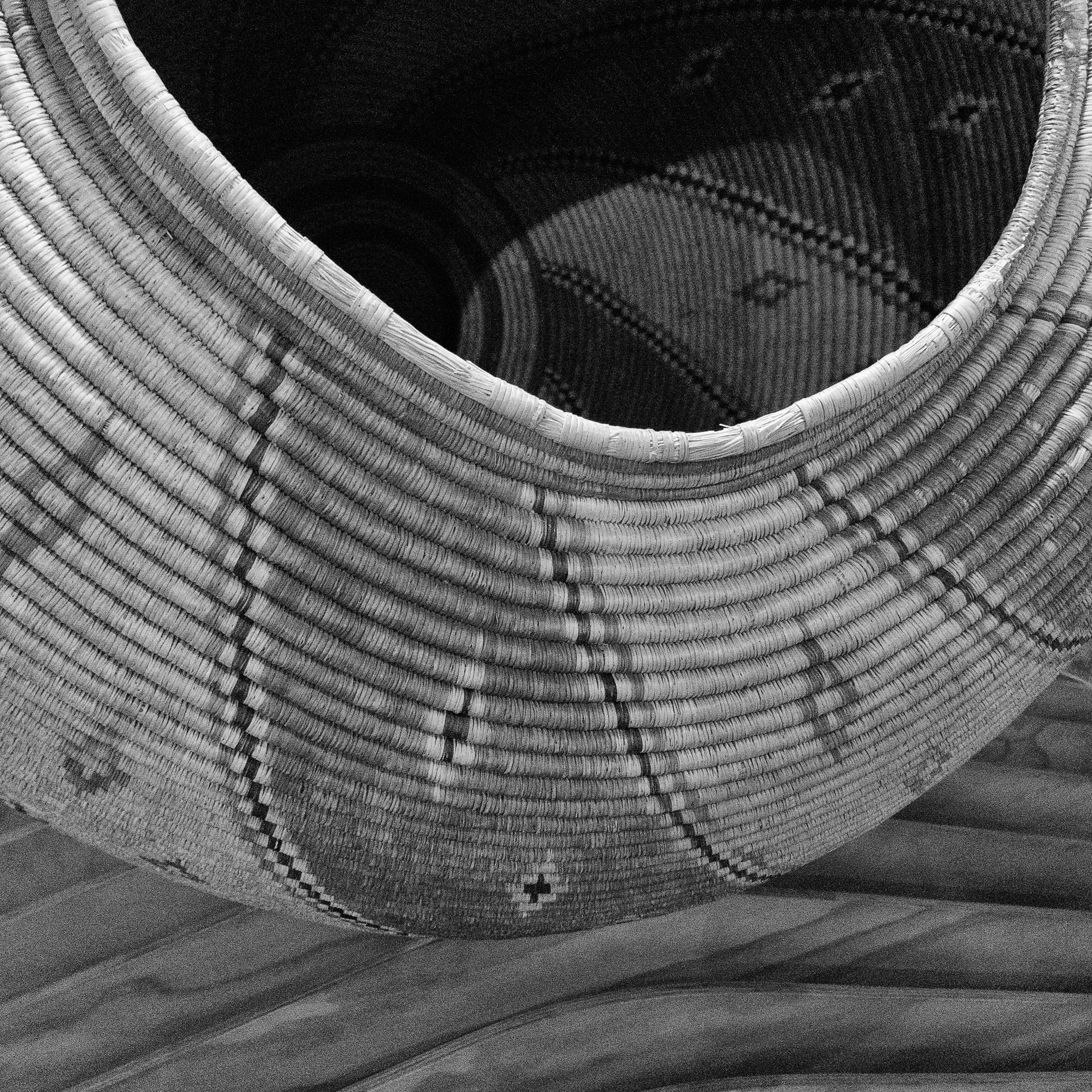 Black and white close-up of a woven basket with intricate patterns, highlighting the texture, curves, and craftsmanship of the handmade fibers.