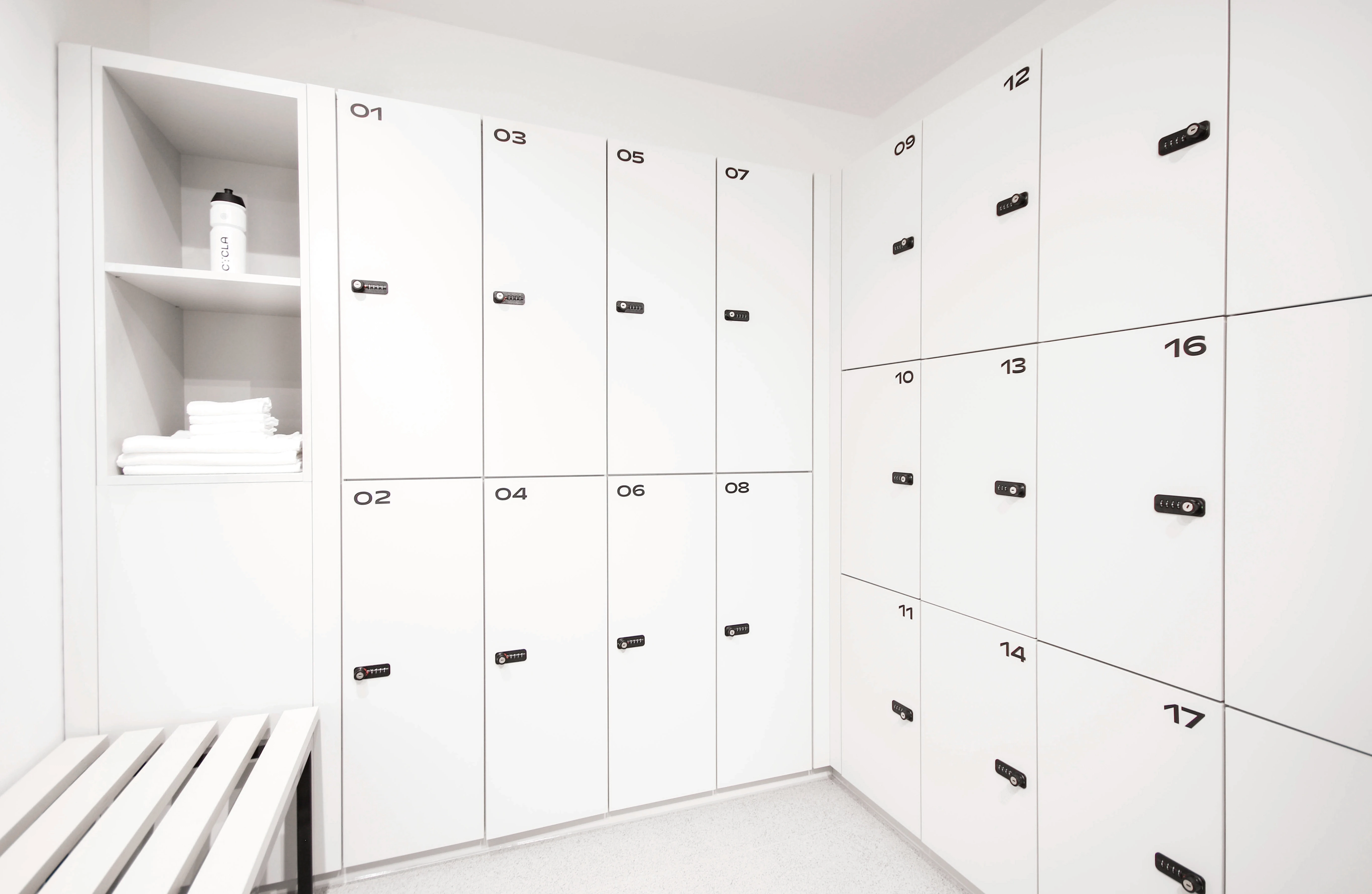 White numbered lockers in the CYCLA changing room with a white slatted bench and an open shelf with towels and a water bottle.