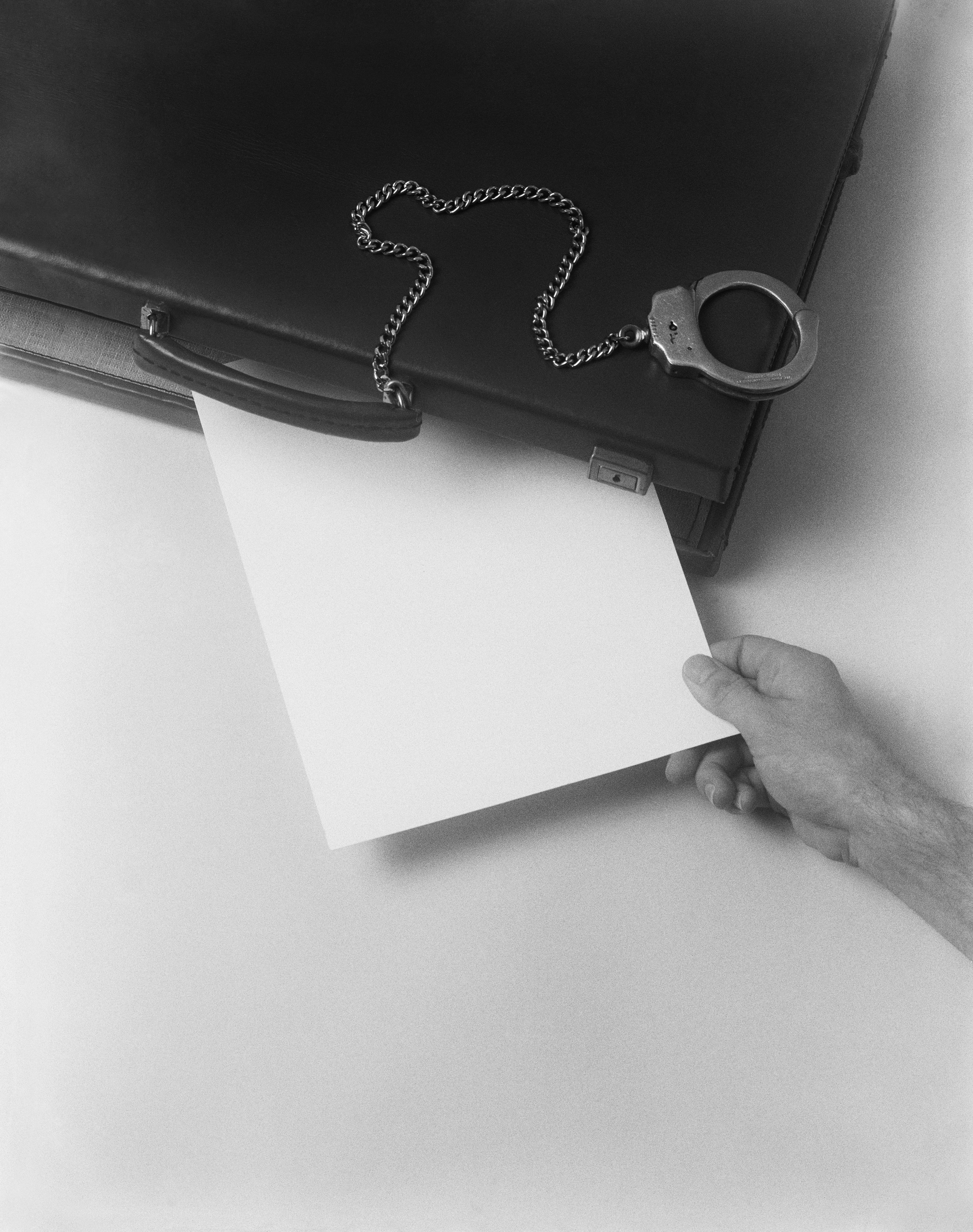 Black and white photograph of a hand pulling a blank sheet of paper from a briefcase with handcuffs and a chain attached, illustrating a binding contract trap.
