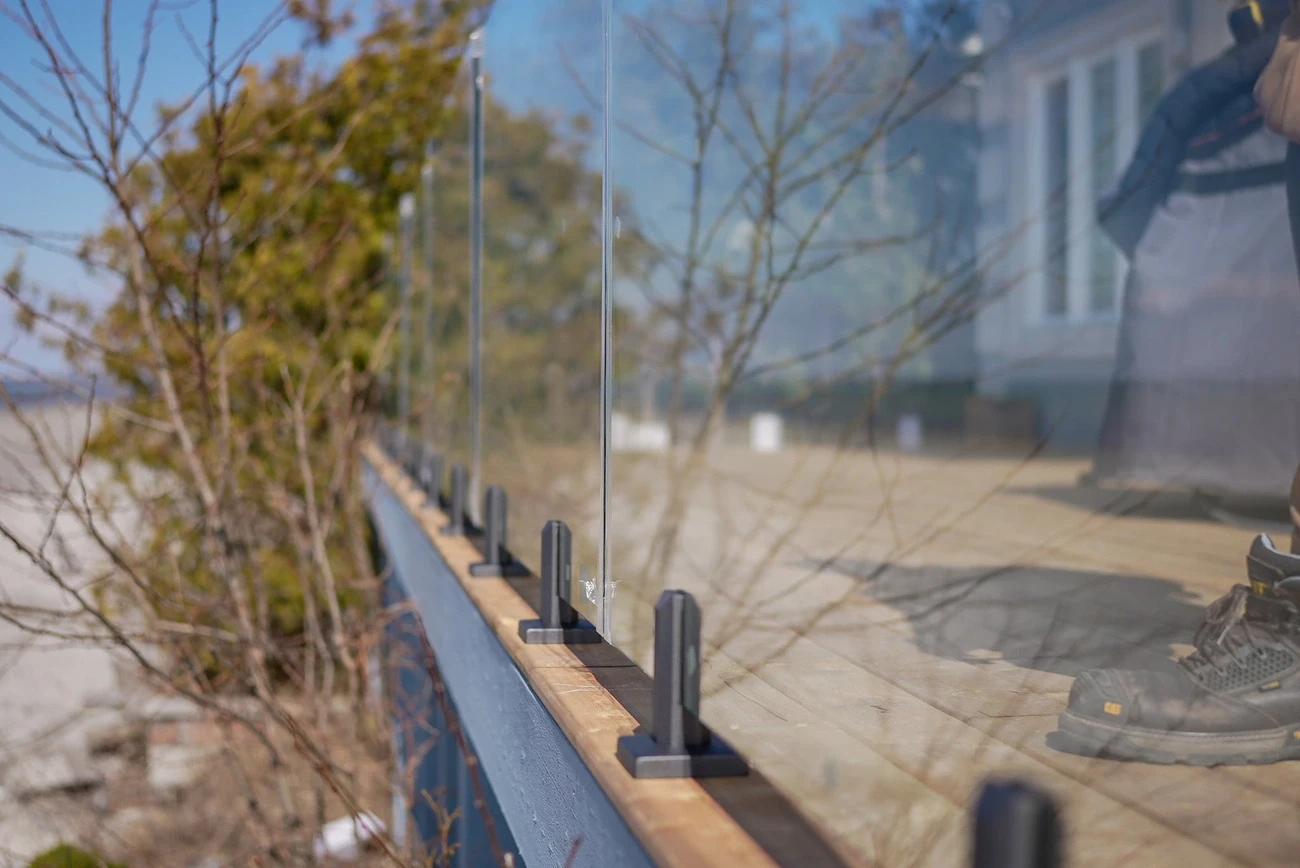 Close-up of a modern glass railing installed on a wooden deck with metal supports and a person standing nearby.