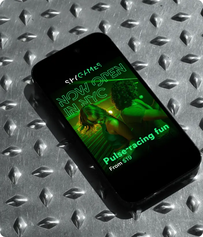 Smartphone on a metallic surface displaying a SPYSCAPE ad with a neon green NYC launch graphic and people taking part in a sensory game.