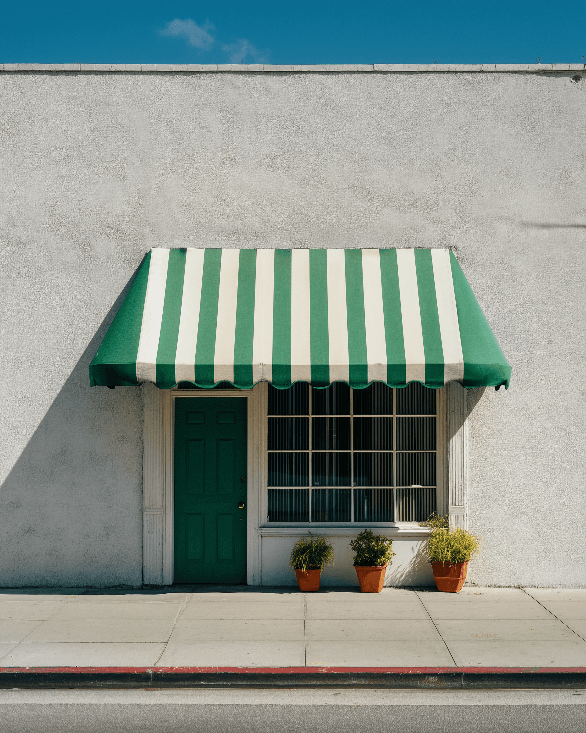 Minimalist facade with red window and striped awning on a white stucco wall under a clear blue sky.