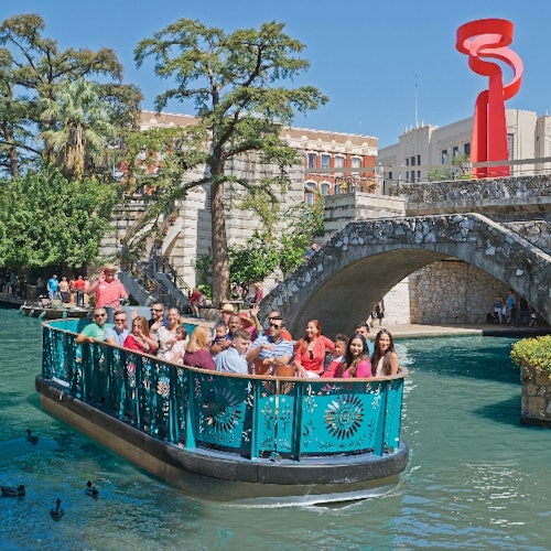 A group of people on a turquoise boat cruise along a canal, with a stone bridge and red sculpture in the background.