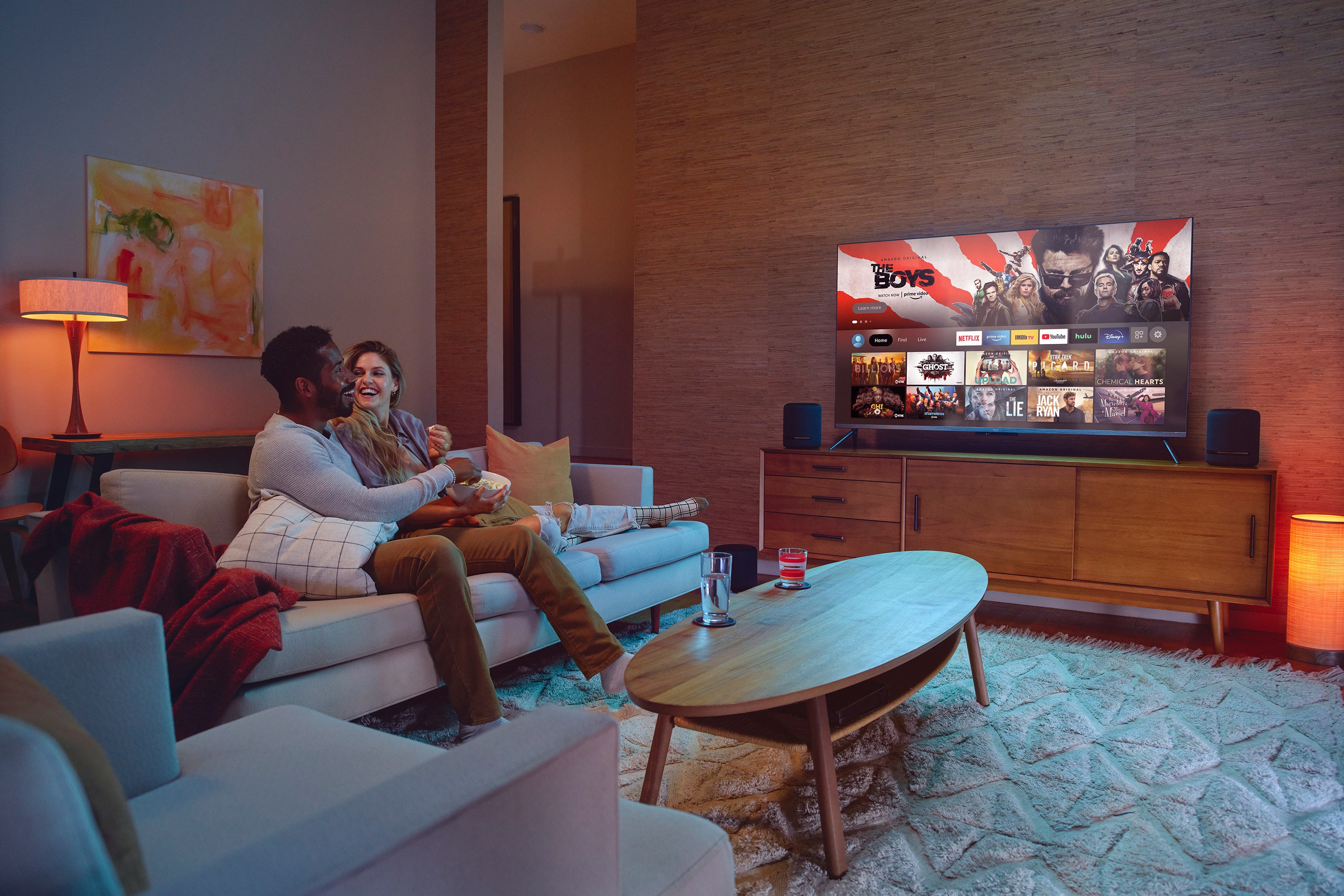 A dimmed living room setting with a male and female sitting closely and looking at eachother while a TV plays on a credenza.