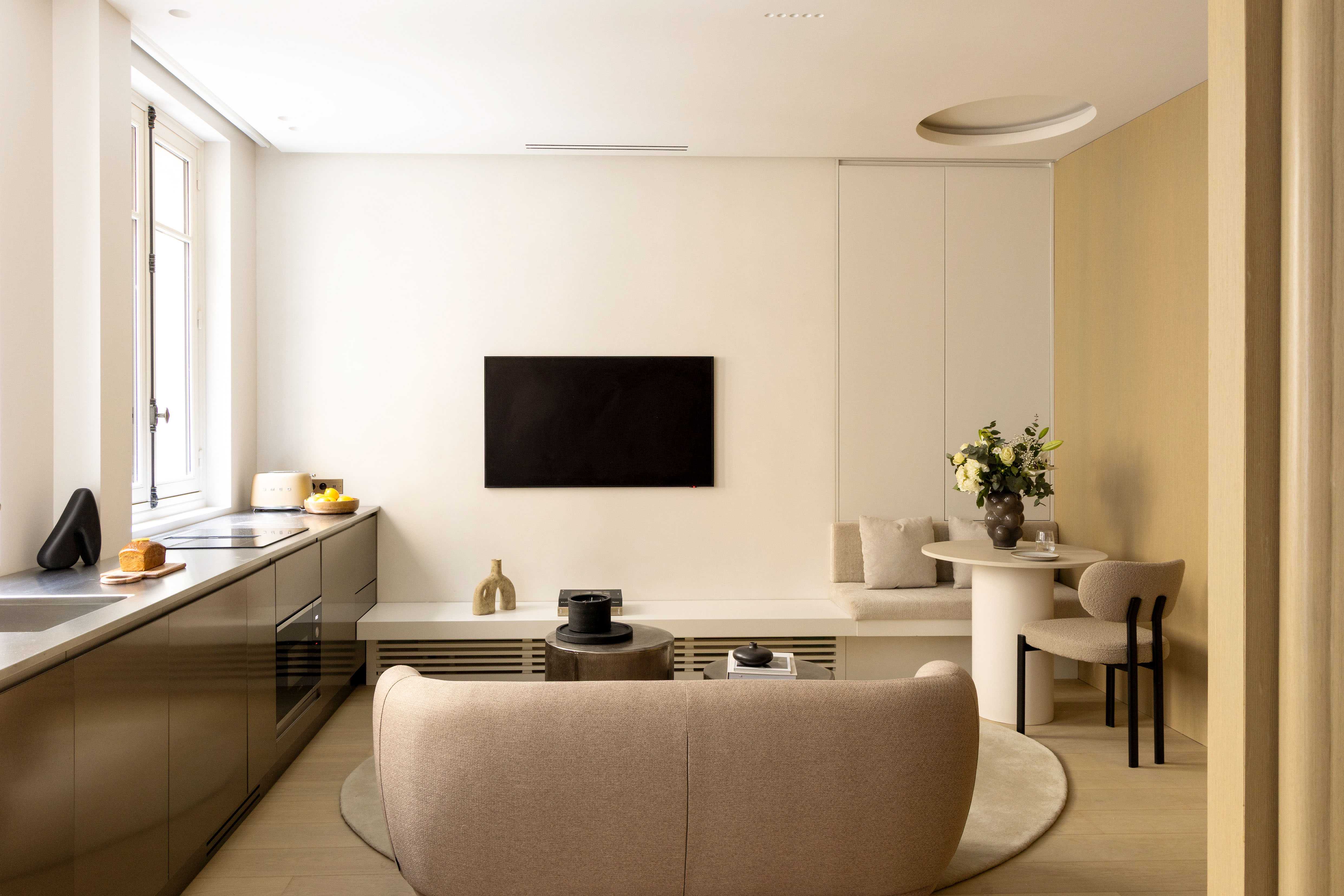 Salon avec canape beige arrondi, TV murale, kitchenette bronze et coin repas en banquette
