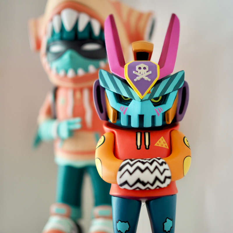 Teq63 collaboration piece by Martian Toys and TenHundred. A mean looking robot figure in an orange hoodie, with a blue mask on. In the background is Hammerhood, another collaboration piece by both artists.