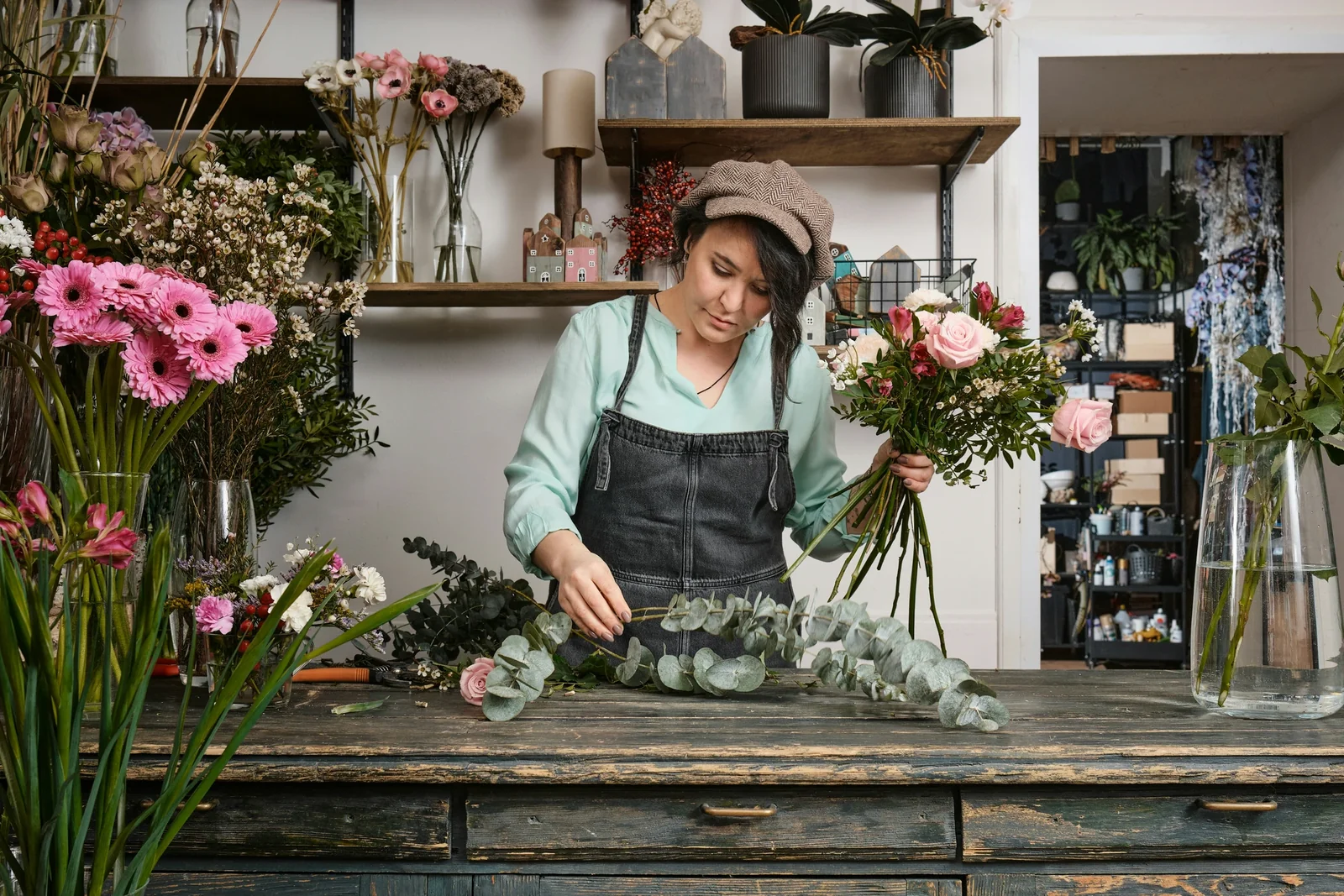 Florist in denim apron arranging pink flowers and eucalyptus at wooden workbench in flower shop.
