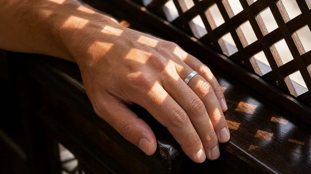 Man's hand wearing a plain silver ring on a dark wooden railing in lattice light