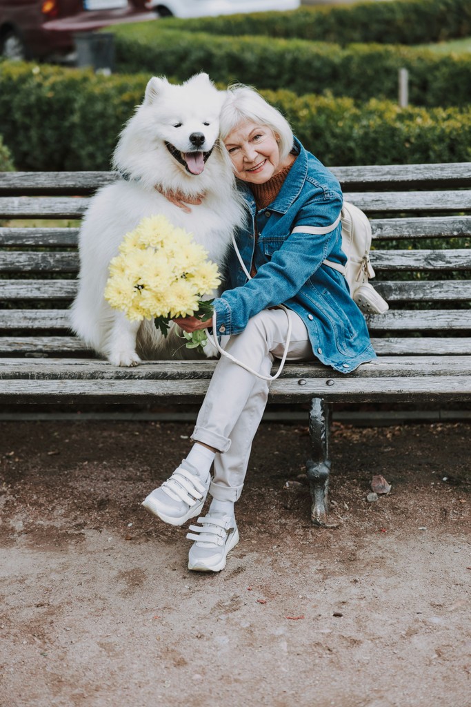 An elderly woman sits on a park bench, joyfully hugging her fluffy white dog while holding a bouquet of yellow flowers, showcasing a heartwarming moment of companionship and love in a serene outdoor setting.