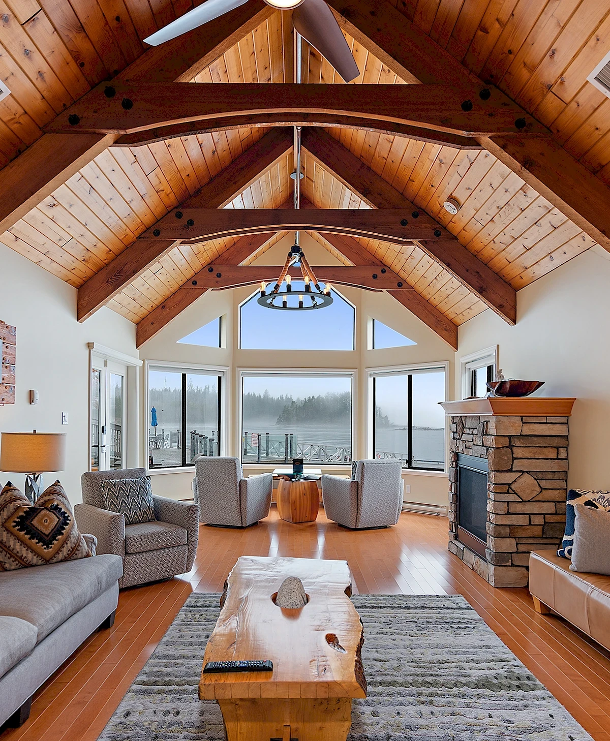 Luxuriöses Wohnzimmer mit hohen Holzdecken, rustikalen Balken, einem Steinkamin und großen Panoramafenstern mit Blick auf eine neblige Küstenlandschaft.