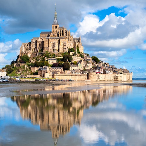 Mont Saint-Michel with its abbey, surrounded by water and reflecting in the stillness, under a partly cloudy blue sky.