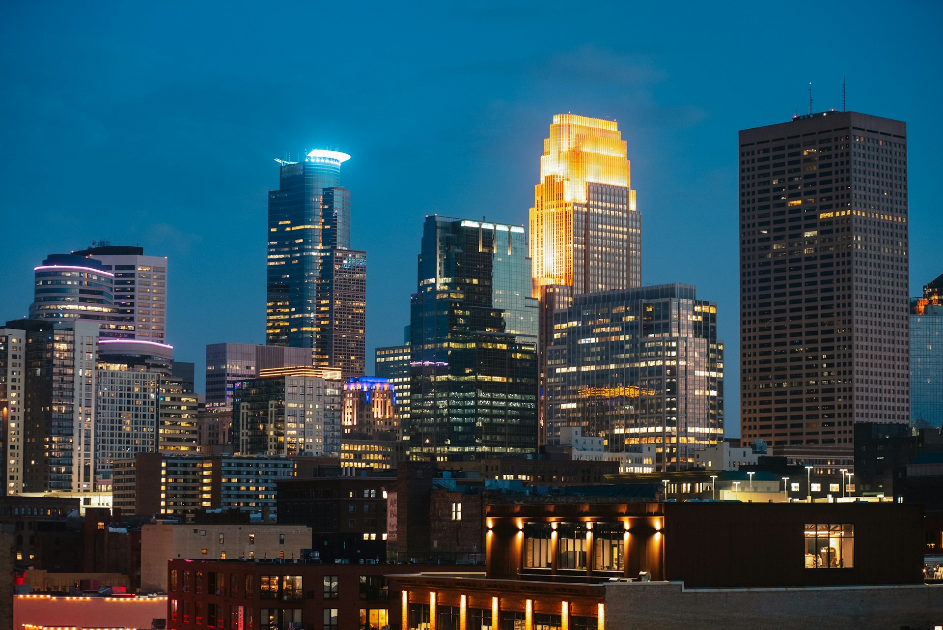 Dense downtown skyline with illuminated skyscrapers glowing against a deep blue evening sky.