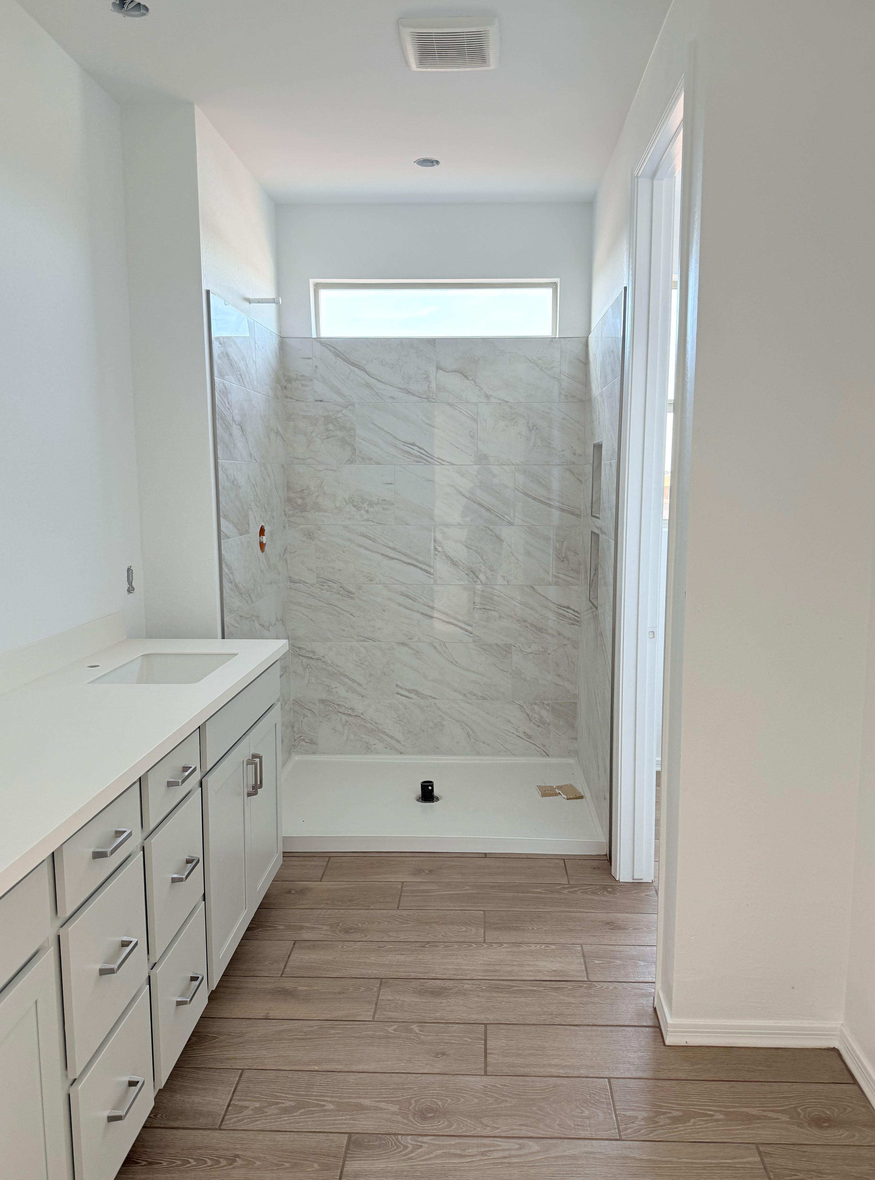 White marble-look tile shower installation with large format tiles and a built-in curb, paired with wood-look tile flooring and a white vanity.