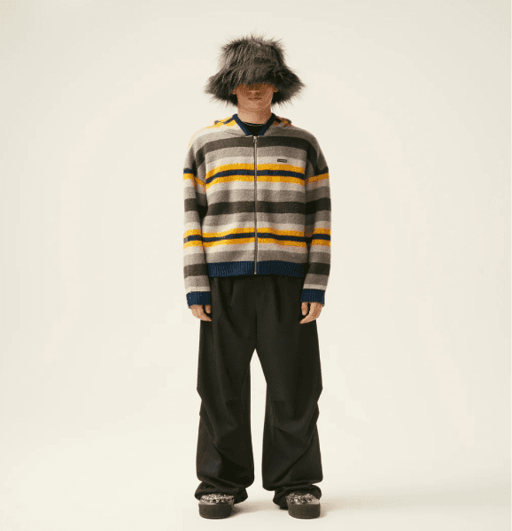 "Male model wearing a striped zip-up sweater with yellow and gray tones, black wide-leg pants, a fur bucket hat, and chunky platform shoes. Unique blend of contemporary streetwear and eclectic fashion."
